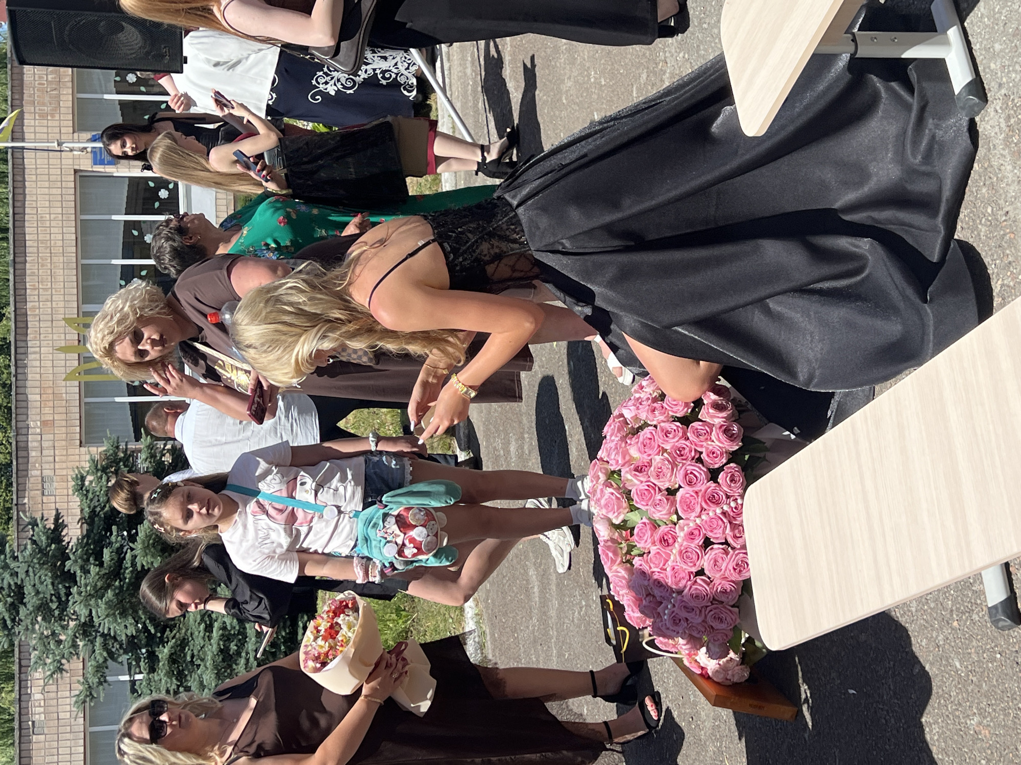 The image captures an outdoor gathering, likely a school event or celebration given the presence of young people in festive attire. Several individuals are visible, including what appears to be a young woman in a formal black dress, possibly a graduate. Other people are dressed in casual clothing, with some holding flowers and bouquets. A large arrangement of pink roses is a prominent object in the scene. The setting seems to be a schoolyard or a public space with a brick building in the background and trees suggesting it's daytime. The lighting indicates a sunny day. The overall mood appears celebratory and happy. No visible text is present in the image.