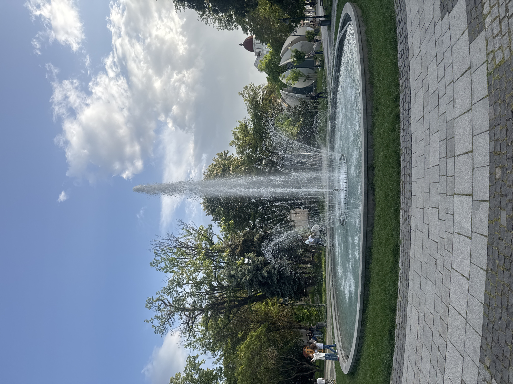 The image shows a large, round fountain in a park. The fountain is actively spraying water high into the air. The sky is partly cloudy with blue patches. Trees surround the fountain, and a building with a red dome can be seen in the background. A paved path runs alongside the fountain, and there are people walking near the fountain. The scene is consistent with being located in the Central Park in Plovdiv, Bulgaria.