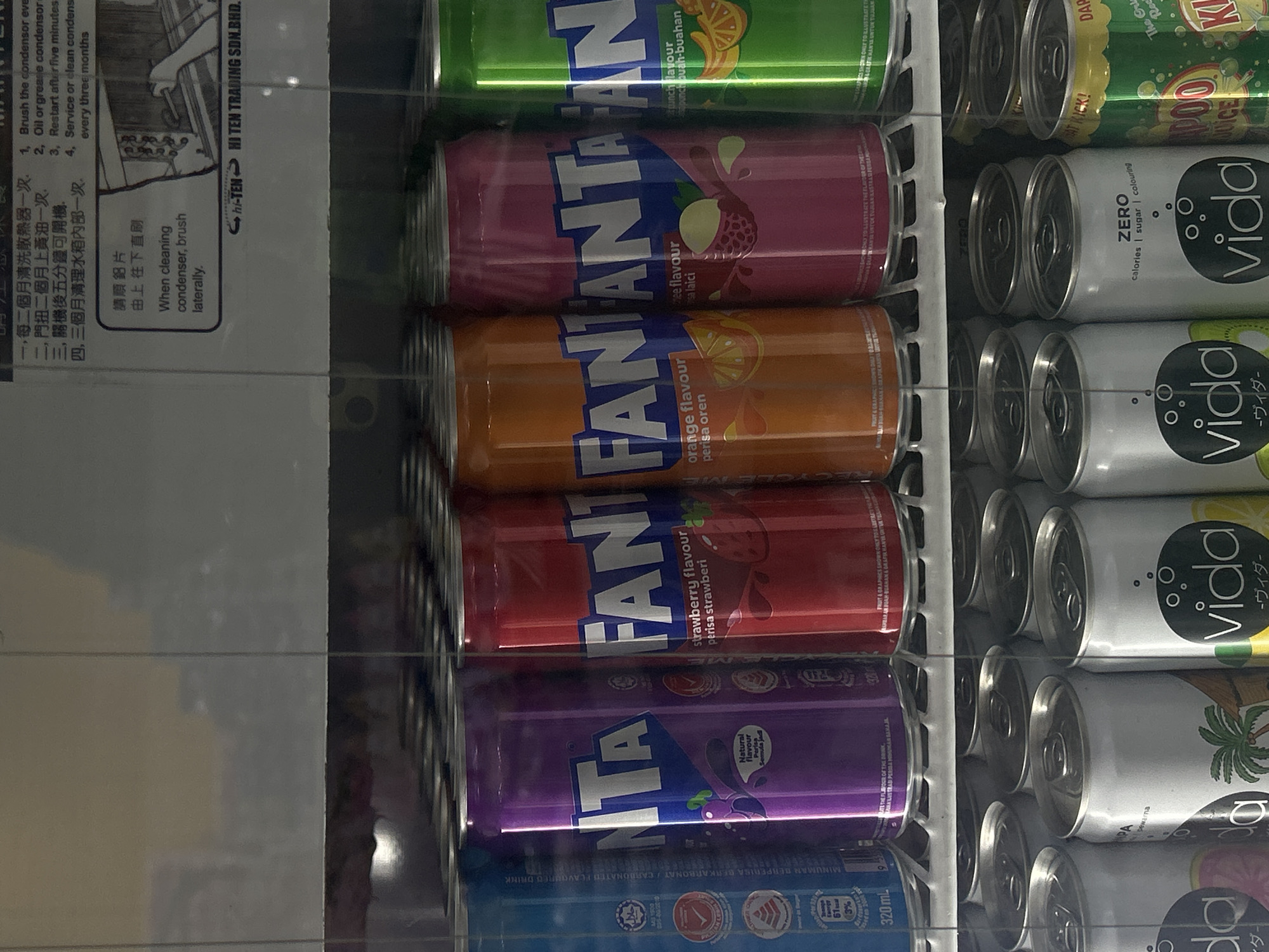 The image shows a refrigerated shelf in Kuala Lumpur, Malaysia, stocked with cans of soft drinks. On the left is a service advisory sticker in Chinese and English. The soft drinks are arranged in columns, with Fanta in a variety of flavors including orange, strawberry, grape, lychee and punch on the left and Vida brand drinks on the right.