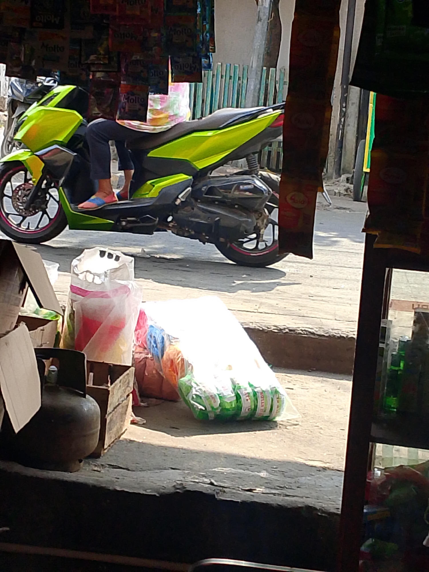 The image depicts a street scene in Palembang, Indonesia, likely in a market or roadside stall area. In the foreground, a large, dark gas cylinder is visible to the left. Next to it are stacked cardboard boxes, one of which is open. To the right of the boxes, several large, clear plastic bags are filled with bundled items that appear to be green bottles with white labels.
Behind these items, a bright lime-green motorcycle is parked. A person is seated on the motorcycle, wearing dark pants and colorful slip-on shoes. The person's legs are extended, with their feet resting on the ground. The motorcycle has a black seat and silver accents.
In the background, there are more motorcycles parked. Overhead, numerous packets of what appear to be powdered drink or detergent are hanging, displaying various colors and text, including "Molto" branding. A wooden fence with vertical green slats is also visible. The ground is paved with concrete, and shadows indicate a sunny day. The overall impression is a busy, informal commercial setting.