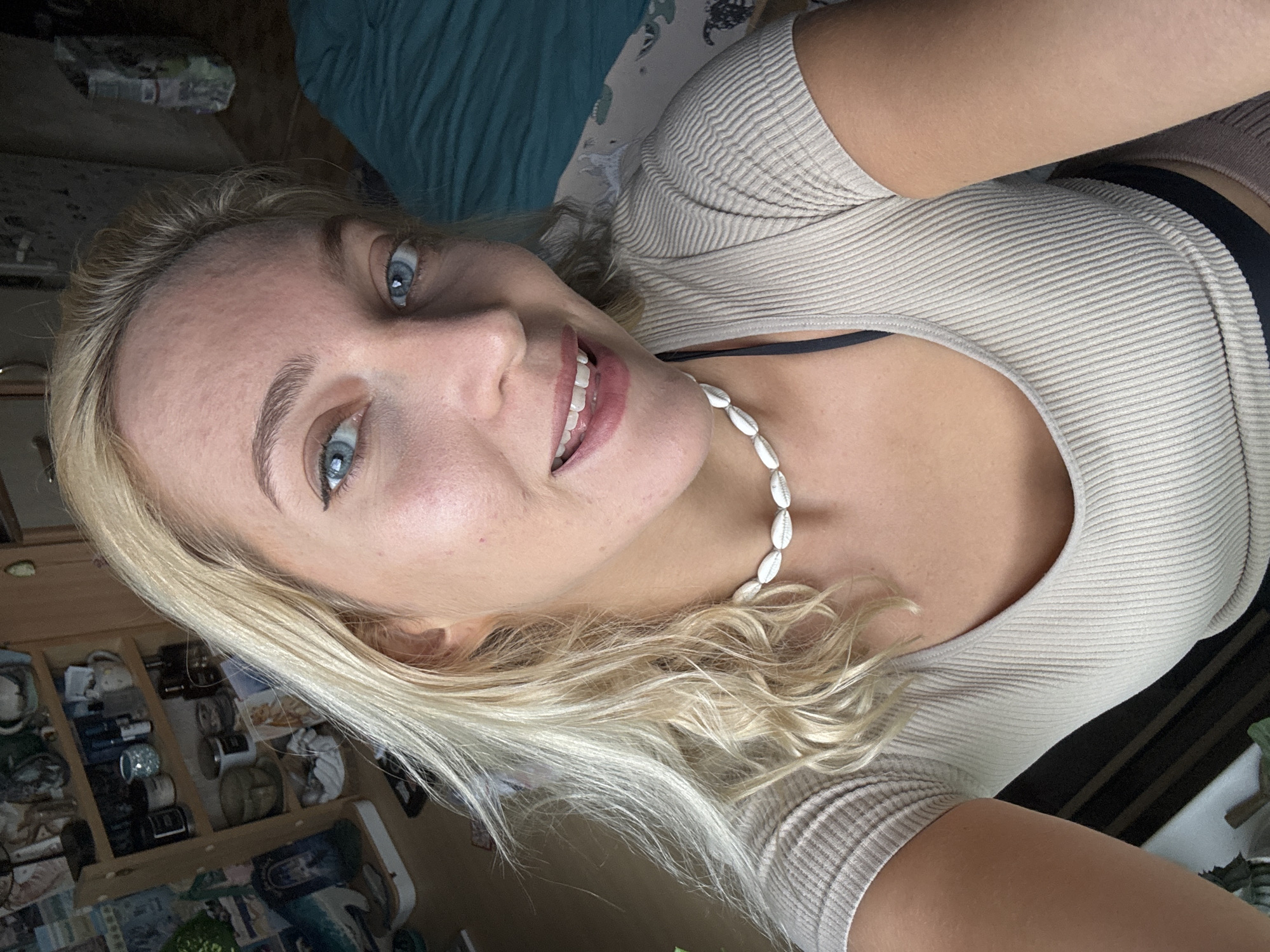 A woman with blonde hair and blue eyes is looking at the camera. She is wearing a beige ribbed top and a necklace made of white shells. Part of a black bra strap is visible. She appears to be indoors, near a wooden dresser filled with various items, and what might be a bed with a blue blanket. There's no evidence to suggest that the picture was taken in the city of Minsk, Belarus.