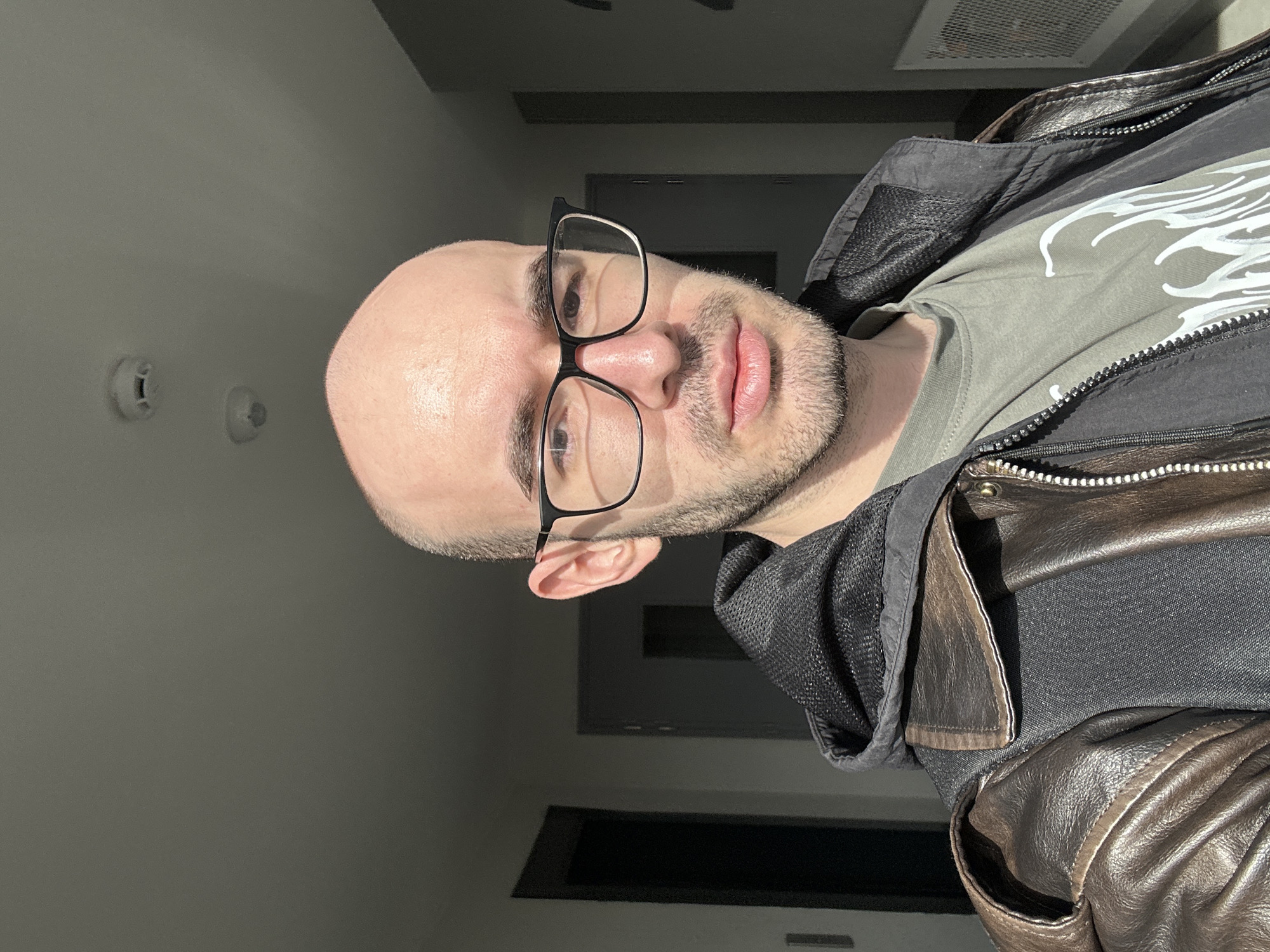 A medium, rotated, indoor shot of a bald man wearing black framed glasses, a green t-shirt, and a leather jacket. The background shows a wall with a couple of plastic wall anchors and an air vent on the ceiling. A doorway is visible in the lower half of the image. The location is not discernible.