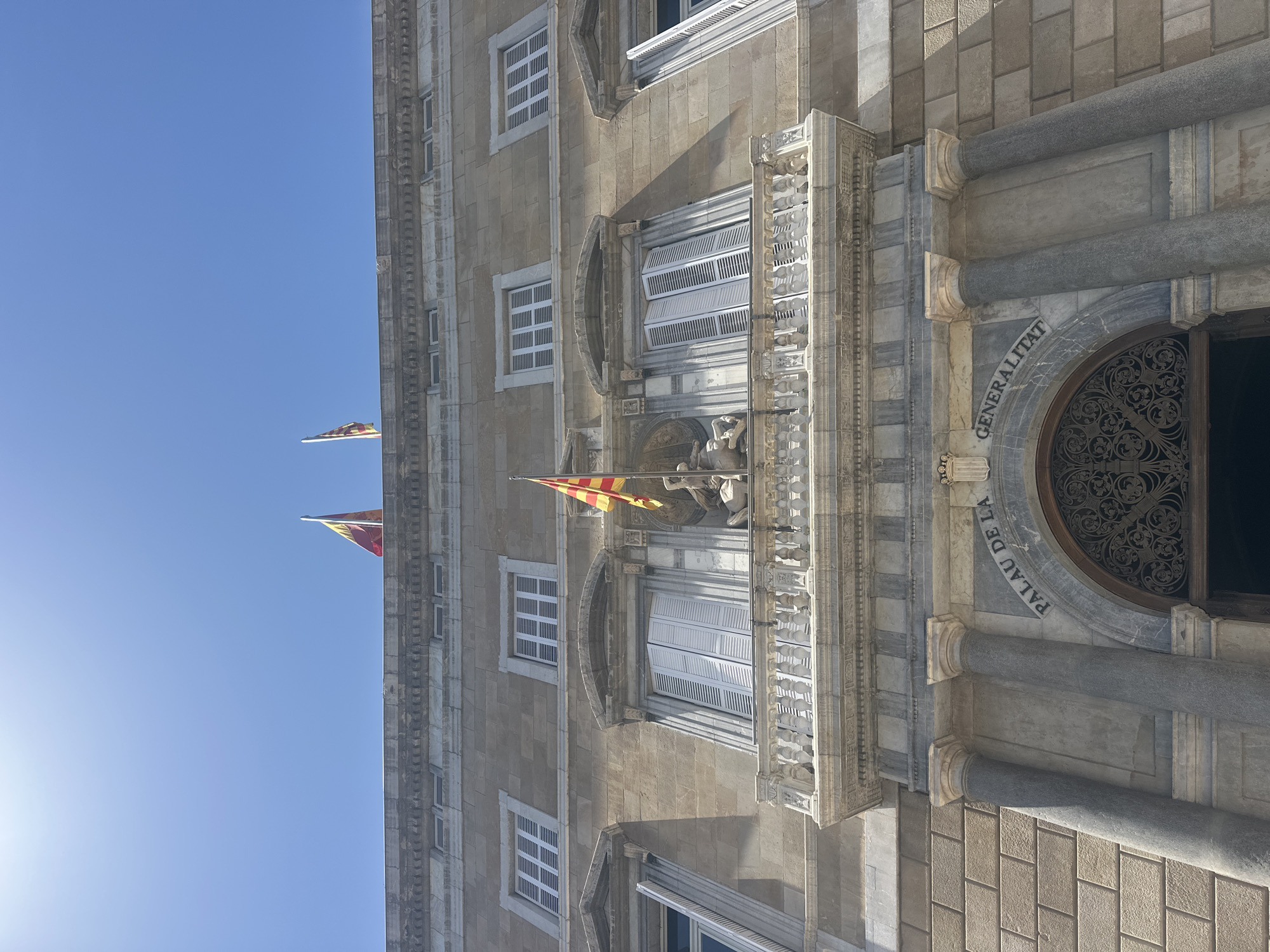 The image captures a low-angle view of the Palau de la Generalitat de Catalunya, a historic building located in the city of Barcelona, Spain. The facade of the building is constructed from stone blocks and features several architectural elements. Dominating the center of the image is an ornate balcony with carved railings, situated above a grand arched entrance. Two large columns flank the entrance, and above the archway, inscribed text reads "PALAU DE LA GENERALITAT". Two Catalan flags, one on the roofline and another on the balcony, are visible, fluttering against a clear blue sky. The windows of the building are adorned with white shutters. A sculpture, partially obscured by a flag, is visible on the balcony. The sunlight casts distinct shadows on the building's facade.