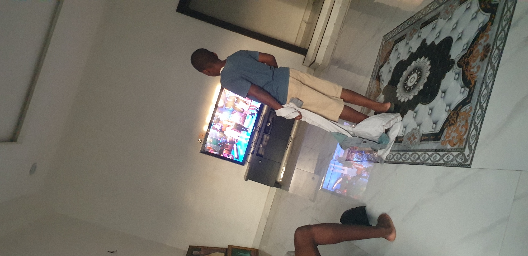 An interior shot captures a living space with predominantly light-colored walls and a highly polished, light-tiled floor reflecting elements of the room. A large, dark-framed flat-screen television is centered on a dark entertainment console, displaying colorful animated or live-action content featuring several human-like figures in what appears to be a stage or performance setting.
In the foreground, a young male, possibly a child or adolescent, stands with his back partially to the camera, facing the television. He has short, dark hair, wears a dark blue t-shirt and light-colored (khaki or beige) shorts, and appears to be wearing dark flip-flops. He holds a bundle of light-colored fabric in his left hand, hanging at his side.
To the far left, the lower legs and bare feet of another individual are visible, suggesting they are seated. A dark bag is positioned on the floor near their feet.
On the floor beneath the standing individual, a rectangular rug with a complex, ornate pattern featuring a central medallion and decorative borders in black, white, and gold/brown hues is laid. The walls are plain, and a recessed light fixture is visible on the ceiling. A dark-framed doorway or architectural feature is present on the right side of the image. On the left wall, two framed pictures are partially visible.
