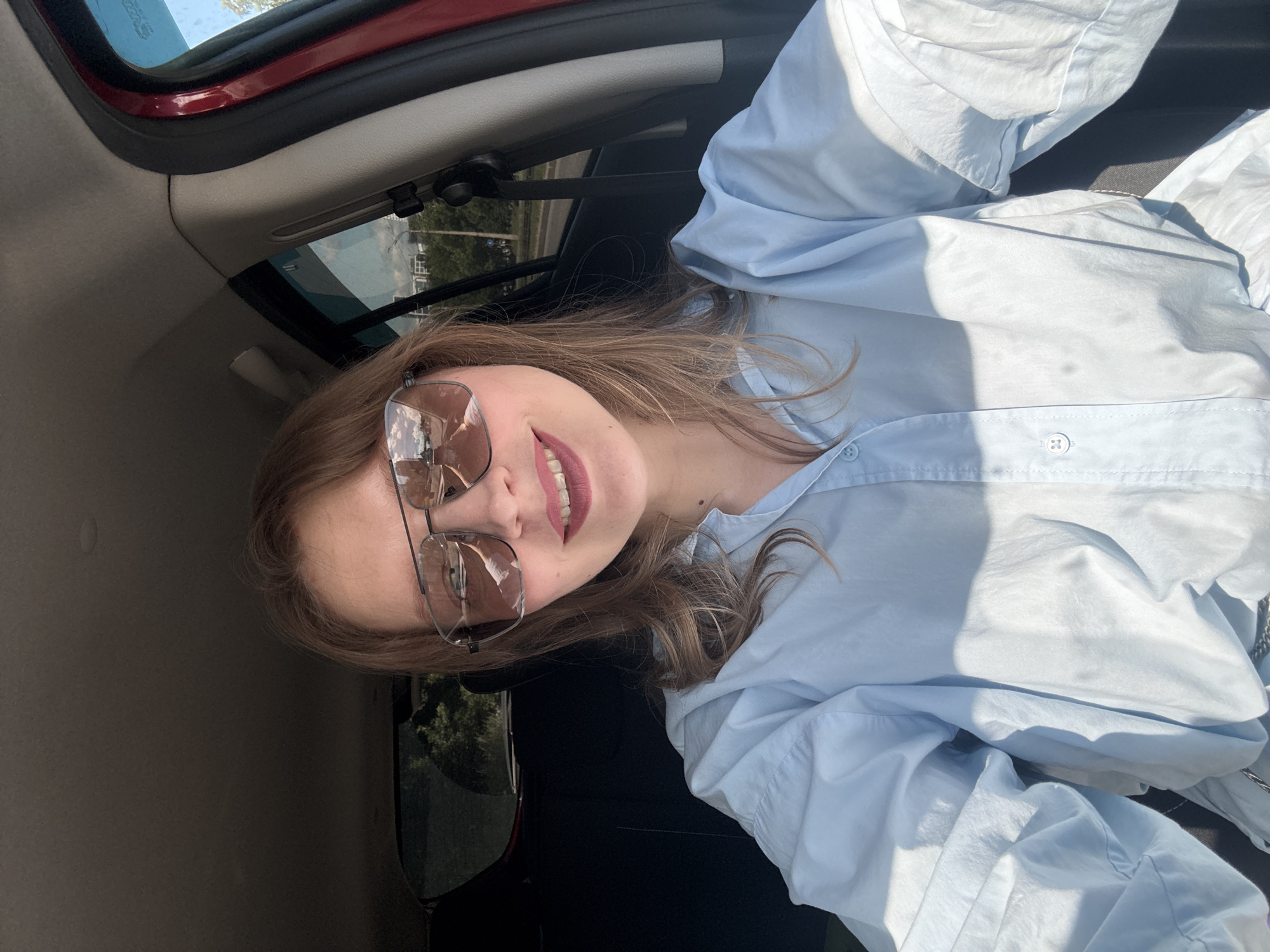 A young woman with light brown hair and fair skin is taking a selfie inside a car. She is wearing oversized aviator sunglasses and a light blue button-down shirt. Her lips are colored with a matte lipstick. The interior of the car appears to be dark, with the red exterior visible on the right side. Sunlight streams in from the right, casting shadows on her shirt. Outside the car window, trees and buildings can be seen, suggesting an urban or suburban setting. The reflection in her sunglasses shows clouds in a blue sky and buildings. The location is identified as Izhevsk, Russia, based on metadata.