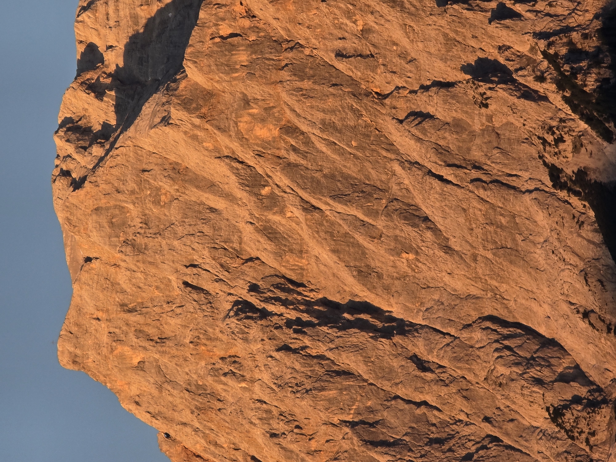 The image shows a close-up view of a rocky mountain face bathed in the warm, orange glow of sunset. The mountain is rugged and textured, with numerous fissures and cracks running down its surface. Shadows are cast in the crevices, accentuating the dramatic topography. The sky above is a clear, pale blue. The cropped images highlight different sections of the mountain, revealing details of its weathered appearance and the interplay of light and shadow. No people, structures, or vegetation are visible in the primary image or crops, suggesting a remote, natural landscape. The location is identified as Lermoos, Austria, which is known for its alpine scenery.