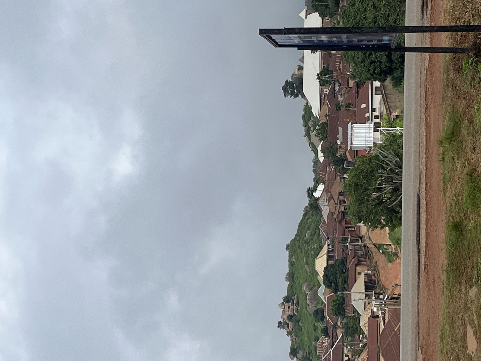 The image captures a panoramic view of a residential area on a hillside in Jos, Nigeria, under an overcast sky. The foreground is dominated by a dirt road leading into the town. A tall, dark signpost stands on the right side of the road, partially obscuring the view.
The hillside is densely populated with houses, characterized by their brown, corrugated iron roofs. Some buildings have a more traditional architectural style, with earthy-colored walls, while others appear to be more modern with white or lighter-colored roofs. Vegetation, including trees and shrubs, is scattered throughout the settlement, with a prominent cluster of trees in the mid-ground.
The upper part of the image reveals a rocky, green-covered hill in the background, dotted with large boulders. The cloudy sky suggests a potentially damp or approaching rainy weather. The overall scene portrays a community nestled into the natural landscape of Jos.