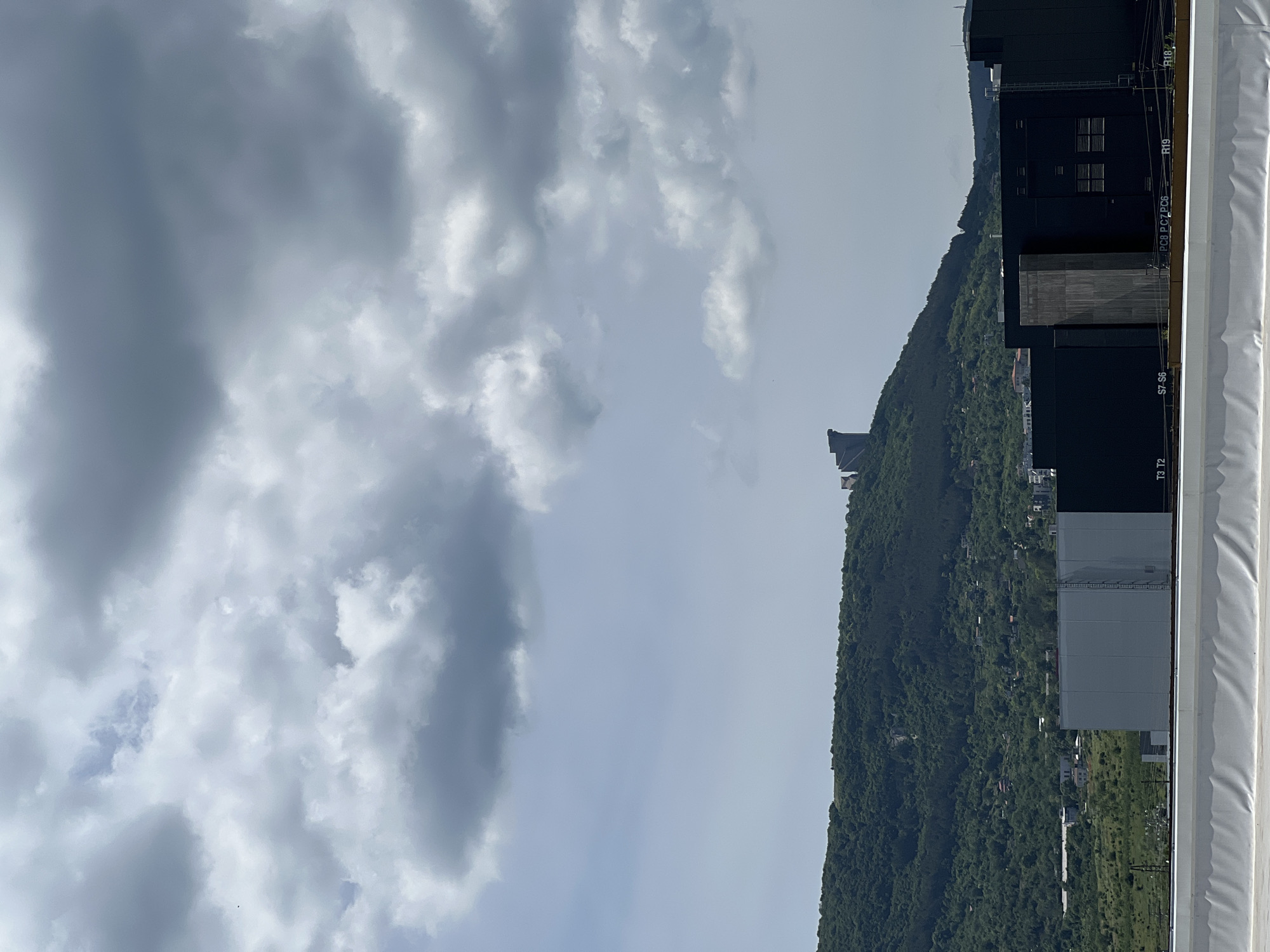 The image captures a broad landscape view under a cloudy sky. The sky, occupying the upper two-thirds of the frame, is filled with a dense cover of grey and white clouds, varying in thickness and density, with some patches of lighter sky visible. Below the cloudy sky, a prominent, verdant mountain or hill range stretches horizontally across the middle of the scene. This range is densely covered in lush green trees and foliage, and several small residential structures are sparsely distributed along its slopes.
In the foreground, occupying the right side of the image, is a large, dark-colored structure, possibly a building or an industrial facility. It appears to be made of dark panels or cladding, with some sections showing small, dark-paned windows. A wide, white railing or barrier runs along the very bottom edge of the frame, suggesting the photo was taken from an elevated position, such as a rooftop or a balcony.
Several alphanumeric labels are visible on the dark foreground structure. From bottom to top, these include "T3 T2," "S7 S6," "P C8 P C7 P C6," "R19," and "R18 Z." The weather appears overcast, and the scene is illuminated by daylight, although the precise time of day is not discernible due to the cloud cover. No human subjects or specific activities are depicted.