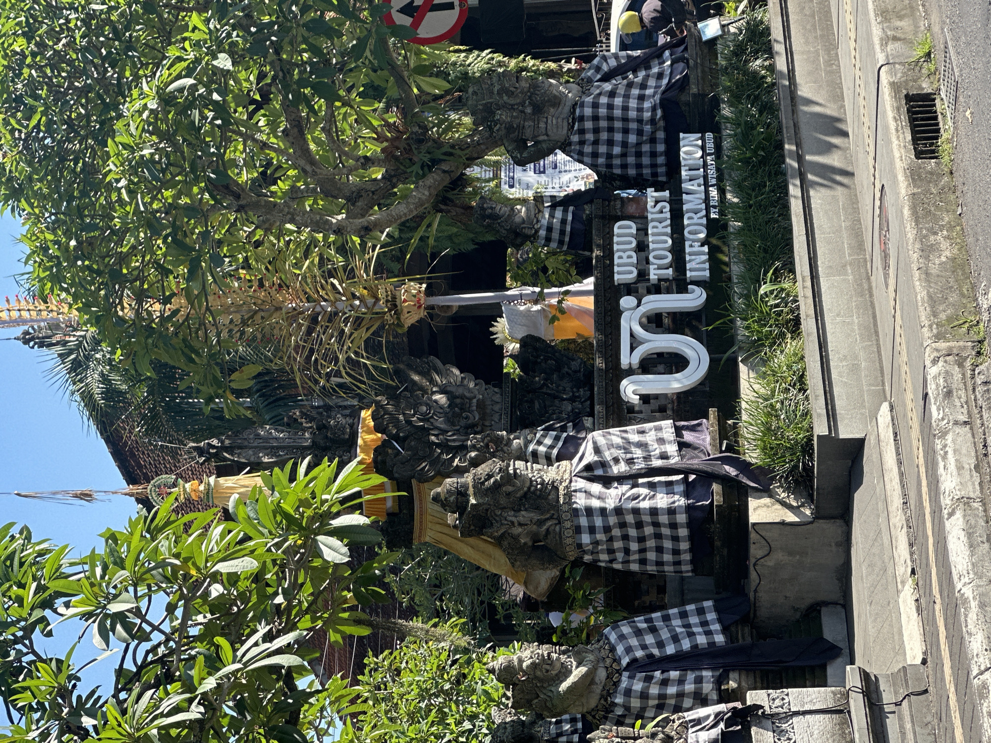 The image depicts the exterior of the Ubud Tourist Information center in Ubud District, Indonesia. Several stone statues of mythical figures, adorned with traditional Balinese black and white checkered sarongs, flank the entrance. A large, white, stylized "UI" logo with the text "UBUD TOURIST INFORMATION" is prominently displayed. Lush green trees and foliage surround the structure, suggesting a tropical environment. In the background, a glimpse of the building's roof and some decorative elements can be seen. The scene is set on a sunny day, with clear blue skies. A traffic sign indicating "no entry" with an arrow is visible on the right side of the image. The foreground shows a sidewalk and a street, with a hint of a motorcycle on the right.