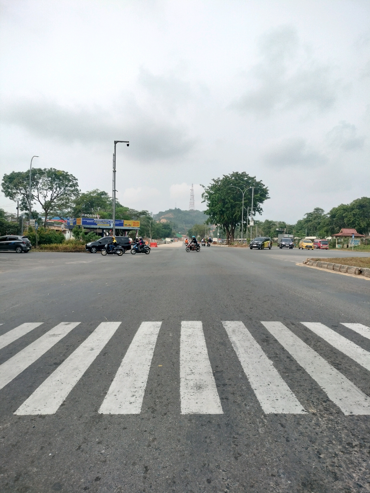 The image depicts a street scene in Batam, Indonesia, with a prominent crosswalk in the foreground. The road is asphalt and appears to be a multi-lane intersection. Several vehicles are present, including cars, motorcycles, and a van. Some people are riding motorcycles, and others are in or near the vehicles. On the left side of the road, there are buildings and trees, with a tall tower visible in the distance. The sky is overcast with clouds. The overall impression is a busy urban environment with moderate traffic.