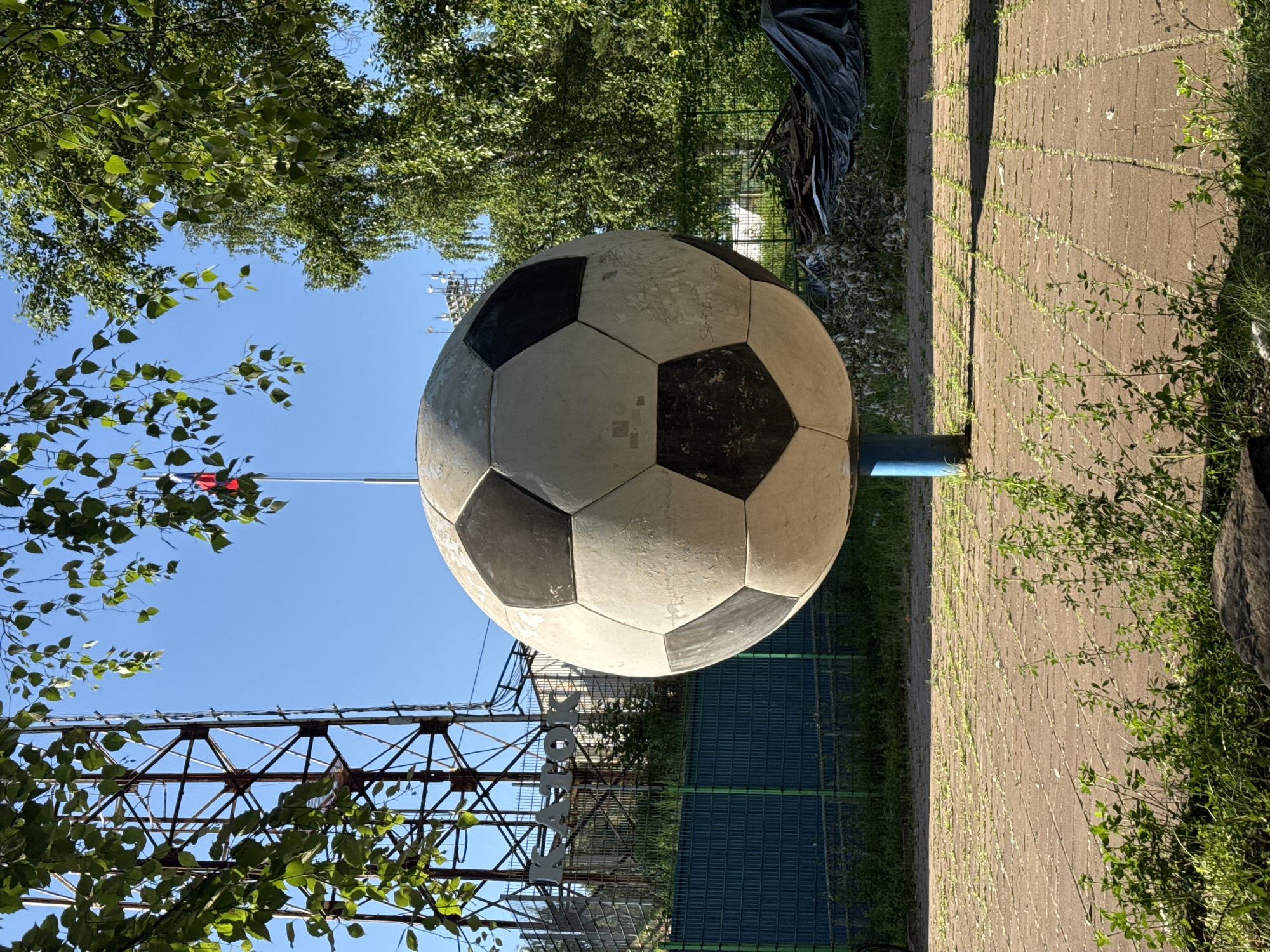 A large, weathered soccer ball sculpture, predominantly white with black hexagonal and pentagonal panels, stands centrally in the frame, mounted on a short blue cylindrical pedestal. The ball shows signs of age, including dirt, scuffs, and some faded graffiti. The ground beneath consists of light brown paving stones, with sparse green weeds growing in the cracks and some thicker green foliage in the immediate foreground.
In the left background, a tall, rusty metal lattice tower, likely a communication or radio tower, dominates the skyline, partially obscured by green leaves from an overhead tree branch. Below the tower, a green mesh fence with solid green panels is visible. Behind this fence, large metallic letters spell out "КАТОК" (KATOK), indicating a skating rink. Further behind the fence are more trees and structures.
In the right background, dense green foliage from trees is prominent. Above the treeline, another, smaller communication tower or mast with multiple antennas is visible. Near the base of the trees, a pile of assorted debris is covered by a dark blue or black tarp.
The sky is clear and bright blue. In the upper right quadrant, a red, white, and blue flag, consistent with the flag of Russia, is visible flying from a flagpole, partially obscured by tree branches. No people are present in the scene.
This scene is located in the city of Izhevsk, Russia, near the Dynamo Stadium, where a similar monument to a football (soccer) ball exists, often associated with adjacent sports facilities, including skating rinks ("Каток").