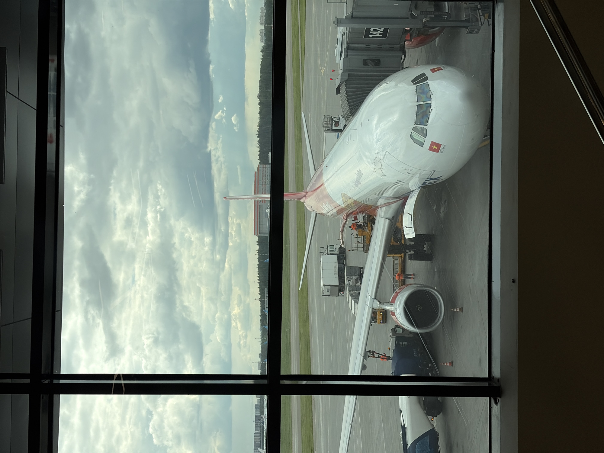 The image depicts a view through a window at Sheremetyevo International Airport, located in Khimki, Russia. An airplane, primarily white with red and gold accents, is connected to gate 142. It features the flag of Vietnam near the front of the aircraft, indicating it is likely operated by Vietnam Airlines. Ground crew personnel in orange vests are working near the engine and under the wing. To the left, the sky is visible, filled with clouds. Outside the window is an airport tarmac and green area.