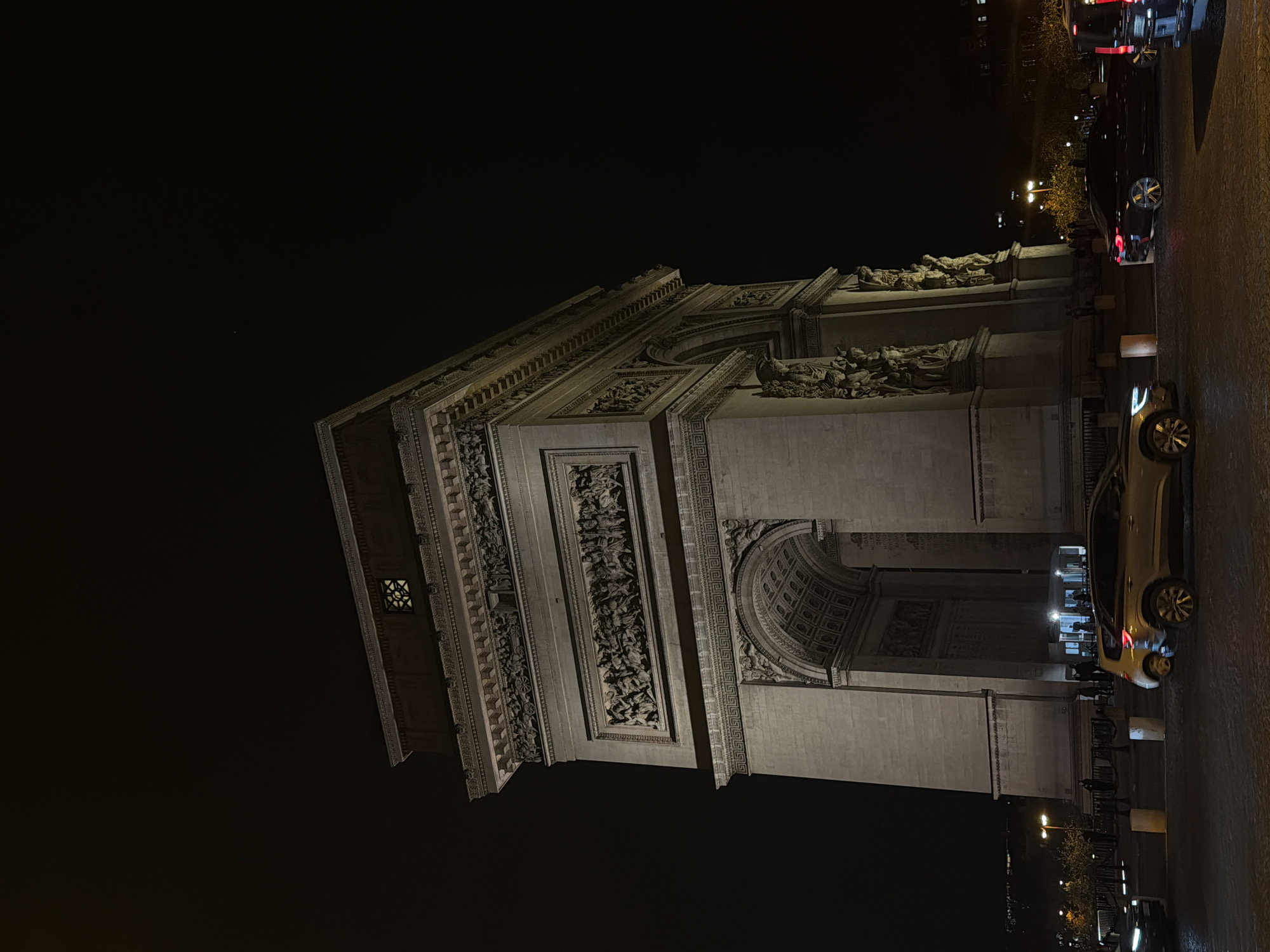 Here is a description of the image:
The image depicts the Arc de Triomphe at night in Paris, France. The monument is brightly lit, showcasing its architectural details, including reliefs and sculptures. Cars are parked along the roadside, and streetlights illuminate the area. People can be seen in the background, suggesting pedestrian traffic. The sky is dark.