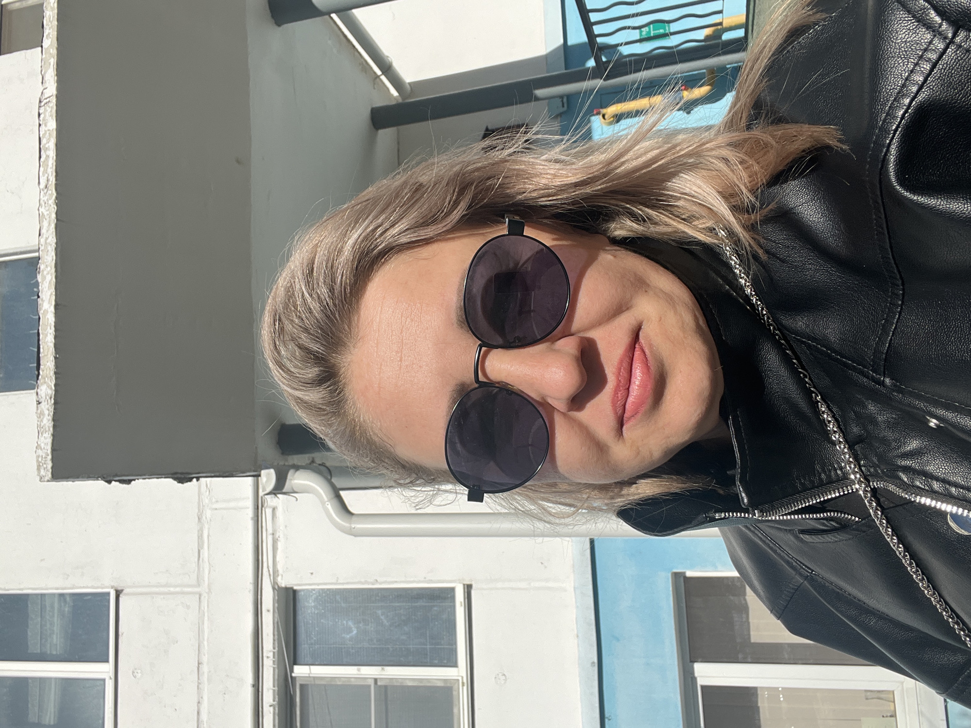 A woman with blond hair, wearing sunglasses and a black leather jacket, poses for a selfie outdoors. The background includes a light-colored building facade with windows and a blue painted section. The location is likely Chaikovsky, Russia.