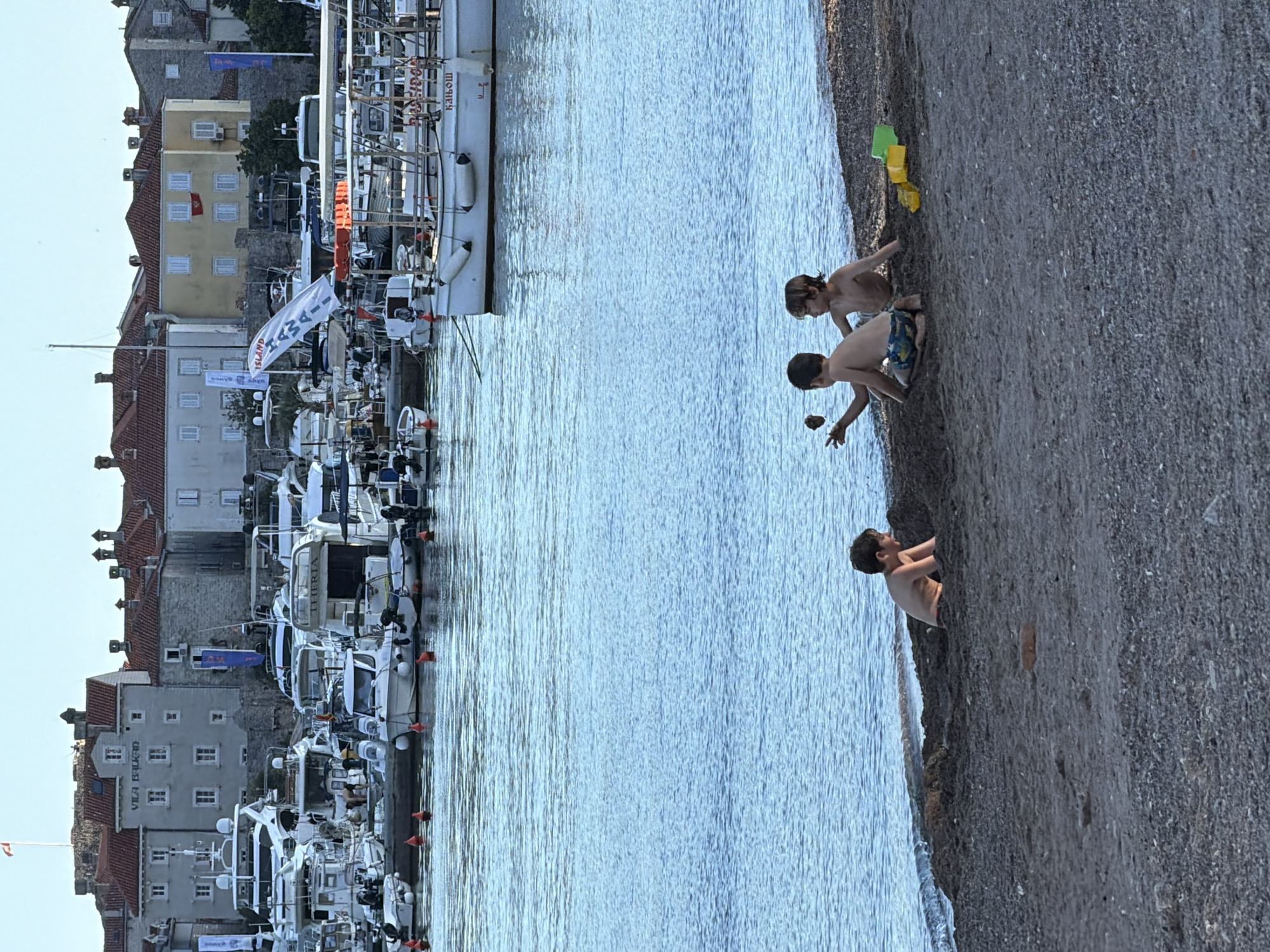 This photograph captures a detailed scene in Budva, Montenegro, specifically near its Old Town and marina.
In the foreground, three young, shirtless boys are playing on a dark-grey pebble beach. The boy on the far left is partially buried in the sand up to his chest, facing right. The middle boy is kneeling, holding a clump of dark earth or sand in his raised left hand, facing towards the boy on the left. He is wearing patterned swim trunks. The boy on the right is seated, facing left towards the other two. Two small plastic beach toys, one yellow and one green, lie on the sand to the right of the rightmost boy. Gentle waves from the clear blue-green sea lap at the edge of the beach.
The midground is dominated by the calm, blue waters of a marina. Numerous boats are docked or anchored, varying in size from small tenders to larger motor yachts. A prominent white tour boat in the central midground displays text including "BASILISK" and "KAIOUW" (likely Cyrillic for "Каиош"), with white fenders hanging from its side. Several orange spherical buoys are visible in the water, indicating mooring points.
The background showcases the historic architecture of Budva's Old Town. Multi-story stone and stucco buildings with red-tiled roofs line the waterfront. A prominent grey building displays "VILA BALKAD" above its windows. A Montenegrin flag (red with a gold emblem) hangs from a yellow building further to the right. Various other flags and banners are visible, including a white banner on a boat or mast reading "ISLAND" and a blue banner on a building. A small red flag atop a fortified structure is visible on the far left horizon. The sky is clear and light blue, suggesting fair weather.