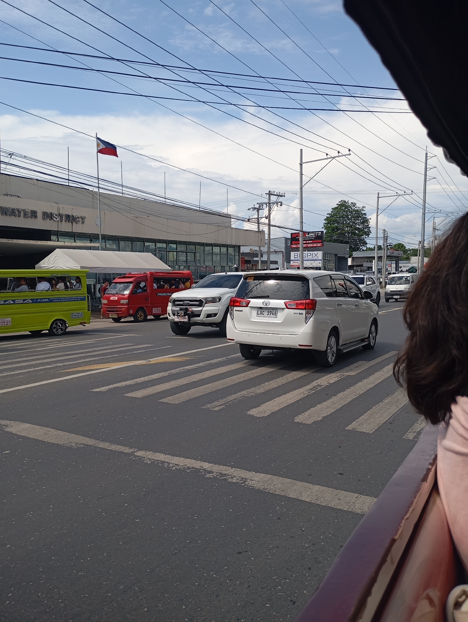 This street-level image captures a busy daytime scene in Davao City, Philippines, from the perspective of a passenger in a moving vehicle, with a person's dark hair visible in the foreground. The weather is bright with a partly cloudy sky.
The main subject is a road with multiple lanes, featuring a zebra crossing. Several vehicles are present, indicative of urban traffic. On the left, a vibrant green jeepney, a common public transport, displays the routes "SASA" and "JP LAUREL" on its side and carries passengers. Adjacent to it is a red passenger van, potentially a multicab or UV Express, with "PANCITAN" written on its side. In the center, a white Ford Ranger pickup truck with a temporary plate "1102-385585" is visible, followed by a white Toyota Innova SUV bearing the license plate "LAC 3946." Other vehicles can be seen further down the road.
In the background, a large building prominently features the text "WATER DISTRICT" on its facade. Above this building, the Philippine flag, red field below the blue, flies on a flagpole against the sky. Numerous utility poles and a dense network of overhead power lines crisscross the scene, a common sight in Philippine cities. Beyond the immediate buildings, a large green tree and additional structures with partially visible signs like "SP LIMITS" and "B_GPIX" are present. The scene depicts a typical urban environment during a regular operating day.