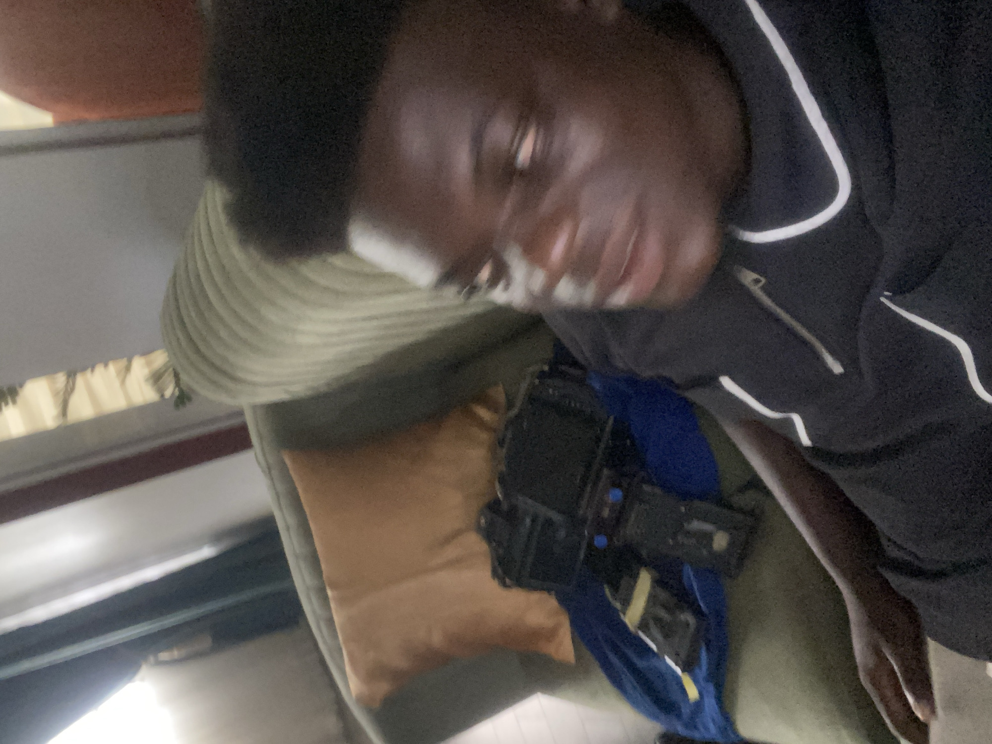 The image displays a close-up, rotated portrait of an individual indoors. The subject is a person with dark skin and short, dark, curly hair, shown from approximately the chest up. They are wearing a black top adorned with prominent white piping along the seams and a zipper on the front. Their face is somewhat blurry, indicating either motion blur or low-light conditions, and they appear to be looking slightly off-camera.
The individual is positioned on soft furnishings, likely a couch or bed. Behind their head is a light-colored, textured cushion, possibly ribbed. Adjacent to them, and supporting their arm, is a plain light brown or tan cushion. On this cushion, next to the person's arm, rests a black and blue electronic device, which appears to be a camera or a similar gadget, with visible buttons and components. The background is dimly lit and indistinct, showing glimpses of interior elements such as a wall or other furniture, and some light sources. No discernible text is visible within the image.