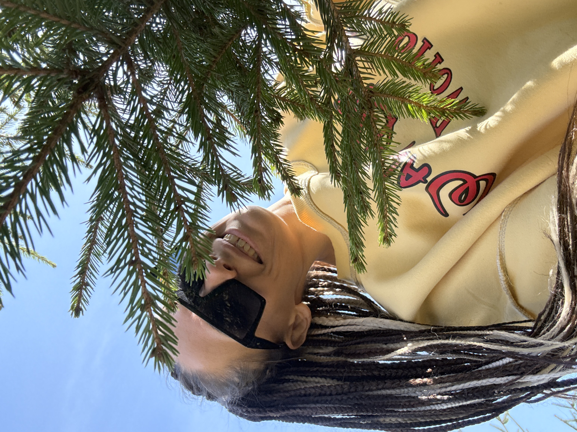 A woman with sunglasses is smiling upward, framed by evergreen branches against a bright blue sky. She is wearing a light yellow hooded sweatshirt with red lettering. Her hair is styled in braids with a mix of dark and light strands. The scene appears to be outdoors. Due to the limited information, the exact location within Yaroslavl', Russia cannot be determined.