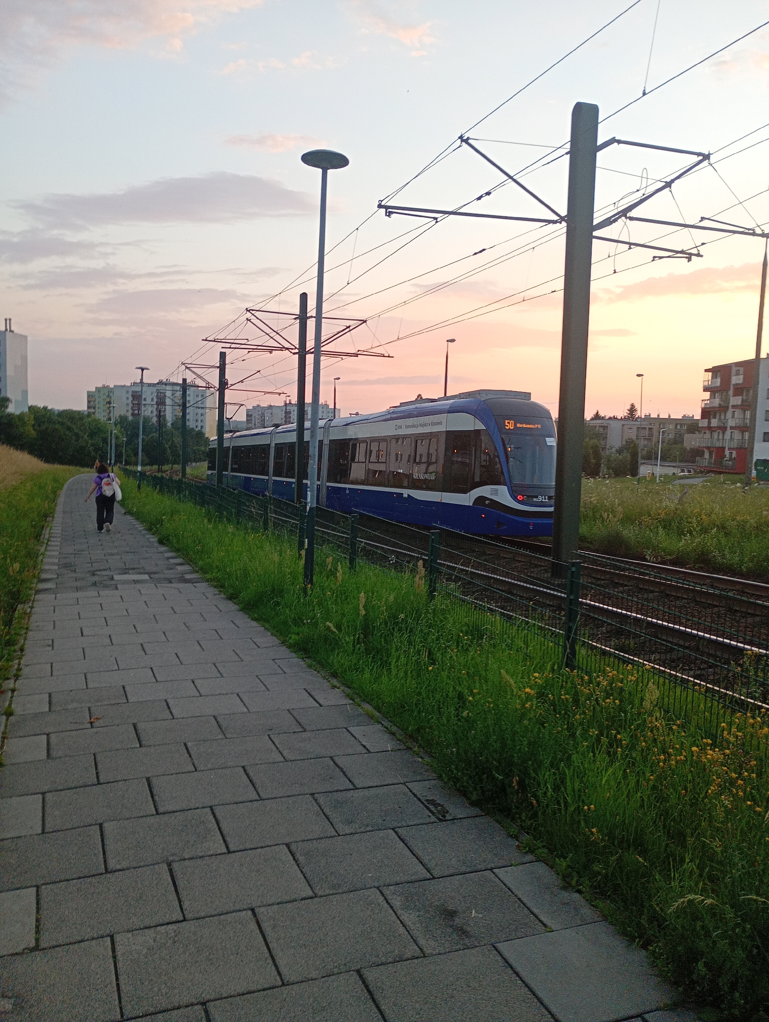 A tram moves along its tracks beside a grassy area and a paved walkway at dusk in Krakow, Poland. The tram, which is predominantly blue and white, displays the number 50 on its front. A person with a backpack walks away from the camera on the paved walkway, heading towards the tram. The sky is filled with pale pink and white clouds, indicative of a sunset. In the background, several apartment buildings and trees line the horizon. Electrical wires for the tram are visible overhead, supported by poles. The overall environment appears to be a suburban or edge-of-city location.