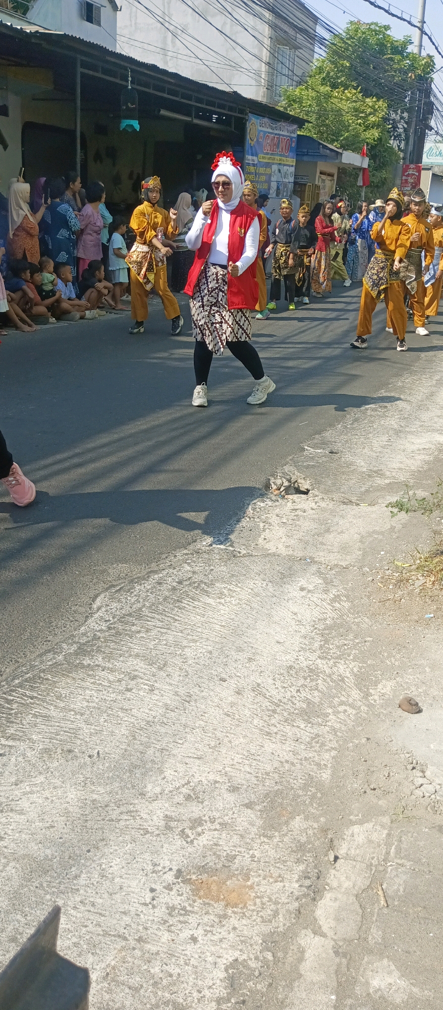 In Malang, Indonesia, a parade is taking place on a street. The foreground is dominated by the rough, textured asphalt of the road, with visible cracks and debris. Shadows of people and objects are cast across the surface. In the midground, a group of individuals are participating in the parade. A woman in the center wears a white hijab, a red vest, and a patterned sarong, and appears to be leading or performing. She is flanked by children and adults dressed in traditional attire, primarily in yellow and brown, with some wearing headbands. The background shows buildings with corrugated metal roofs, overhead wires, and trees, indicating an urban setting. Spectators are visible on the left side of the street, seated and standing along the curb. The overall atmosphere suggests a cultural event or celebration.