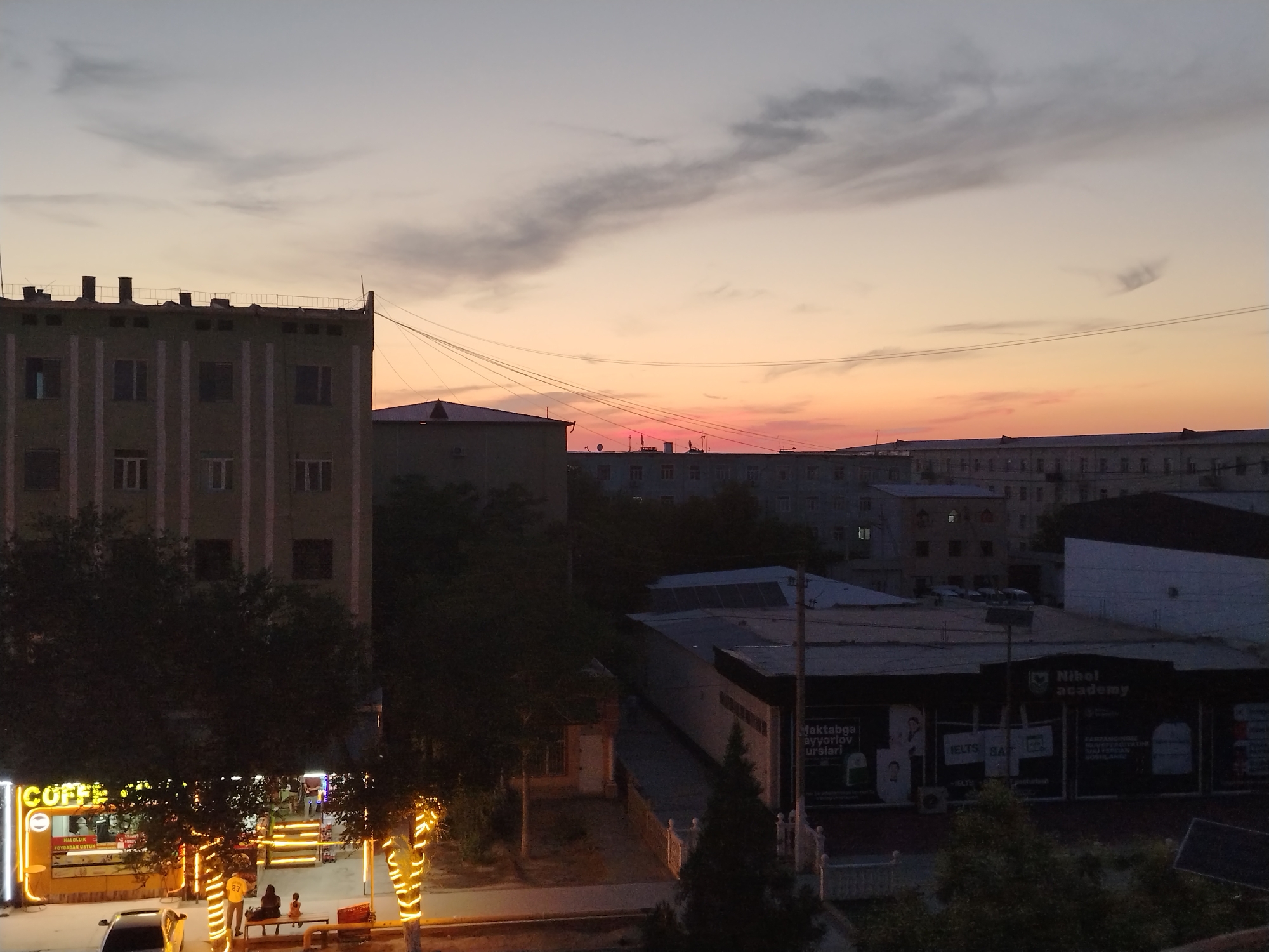 The image shows a cityscape at dusk, with a gradient of sky colors ranging from pale gray to orange and pink near the horizon. In the foreground, on the left, there is a brightly lit storefront with "COFFE" prominently displayed in yellow neon lights. The storefront appears to be a cafe or food establishment, with people visible inside and a few people sitting on a bench outside. Trees decorated with yellow lights line the sidewalk in front of the storefront. A car is parked on the street to the left of the cafe.
Behind the cafe, there are several buildings of varying heights. To the left, a large, multi-story building with a flat roof and visible chimneys stands out. In the middle ground, a lower-lying building with a dark roof and some lighting is visible. Further back, a long, multi-story building stretches across the frame, with multiple windows. A sign for "Nihol academy" is visible on one of the buildings in the middle ground. The overall scene depicts an urban environment, likely residential and commercial, during the evening hours. The presence of the cafe, streetlights, and building lights suggests it's a populated area within the city. The architecture suggests a post-Soviet era style typical of Uzbekistan.