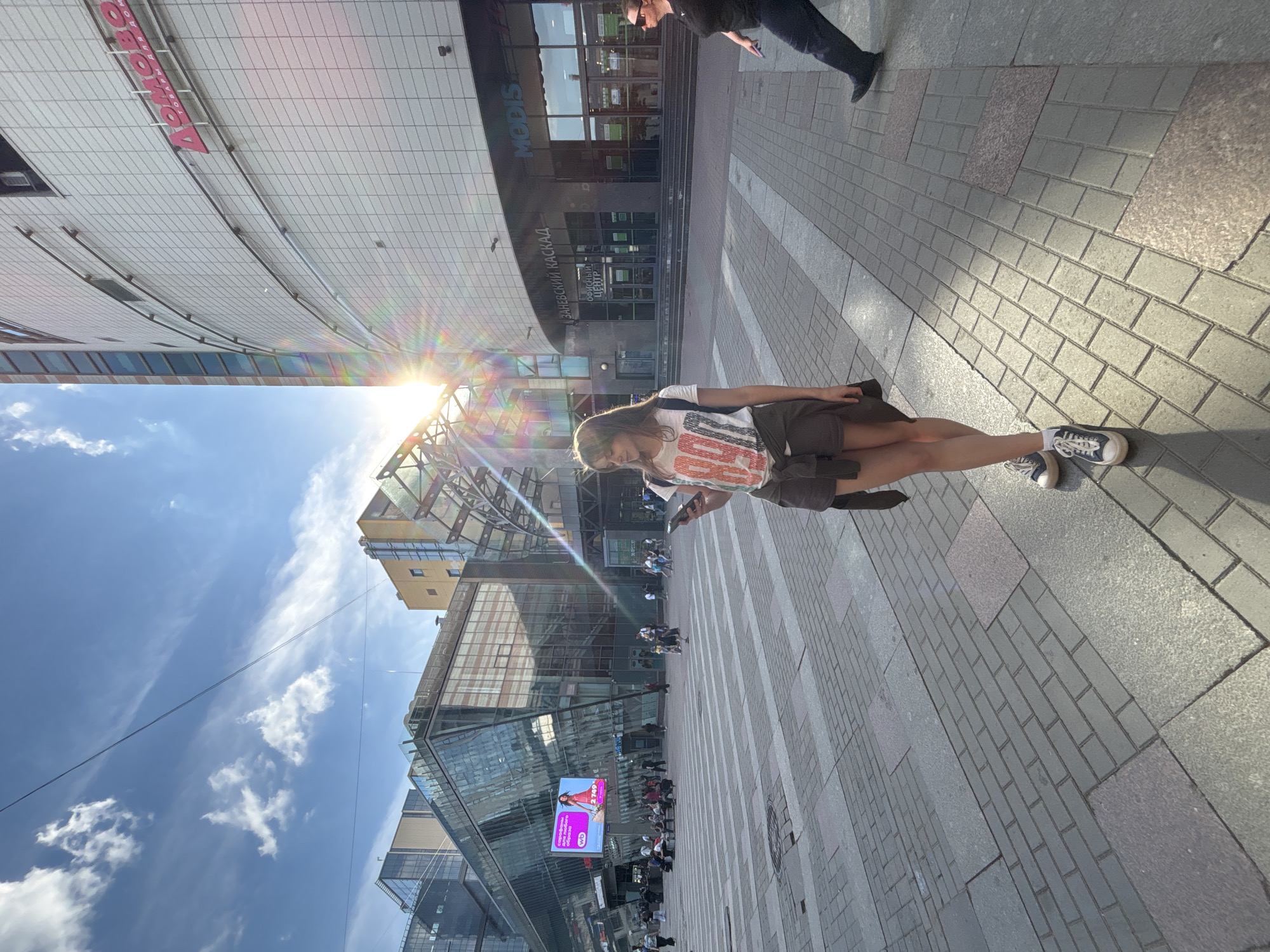 This image depicts an outdoor urban environment during a sunny day, identified as the area surrounding the Zanevsky Kaskad shopping and business complex in Saint Petersburg, Russia. The sky is clear blue with sparse, thin white clouds. Strong sunlight emanates from the upper right, generating significant lens flare and casting sharp shadows across the paved ground.
In the immediate foreground, a young adult female, with light brown hair reaching her shoulders, stands slightly angled to the left, looking down at a dark-colored smartphone she holds with both hands. She wears a white short-sleeved t-shirt with a prominent red, white, and blue graphic, dark brown shorts, and a dark brown jacket or sweater tied around her waist. Her feet are clad in dark blue sneakers with white laces and white socks.
To the far right, a second individual, mostly cropped from the frame, walks leftward. This person, wearing a black top, dark pants, and black shoes, also holds a dark-colored smartphone in their right hand and wears dark sunglasses.
The background is dominated by modern multi-story buildings with extensive glass facades, reflecting the sky and surrounding structures. The large, light-grey building to the right features the Cyrillic signs "ДОМОВО" (Domovo, a home goods store) in red and "MODIS" (a fashion retailer) in dark grey. Further along the building, a sign reads "ЗАНЕВСКИЙ КАСКАД" (Zanevsky Kaskad) and "ОФИСНЫЙ ЦЕНТР" (Office Center). To the left, a glass-fronted building displays a vibrant digital advertisement for "Wildberries" (WB), featuring a woman in a pink dress and Cyrillic text promoting fashion.
The ground is paved with rectangular and square tiles of varying grey and reddish-brown hues, laid in a grid pattern. Numerous other individuals are visible in the mid-ground and background, walking or congregating, indicating an active public space.