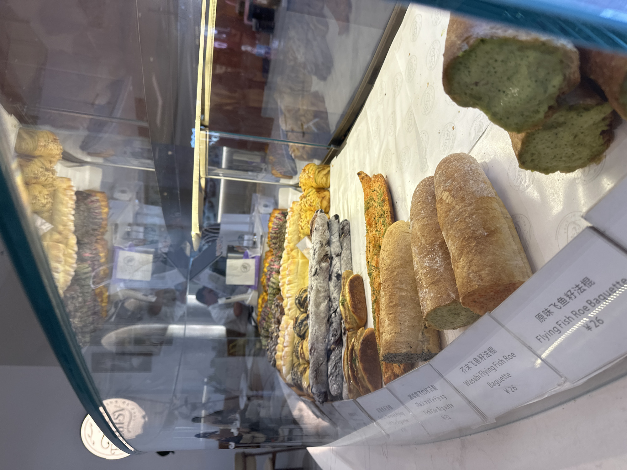 The image captures a display case in a bakery, possibly located in Yuzhong District, China. The case houses a variety of baguettes, each marked with a sign indicating its type and price. The baguettes include options such as Black Truffle Flying Fish Roe Baguette (priced at ¥32), Wasabi Flying Fish Roe Baguette (¥26), and Flying Fish Roe Baguette (¥26).
