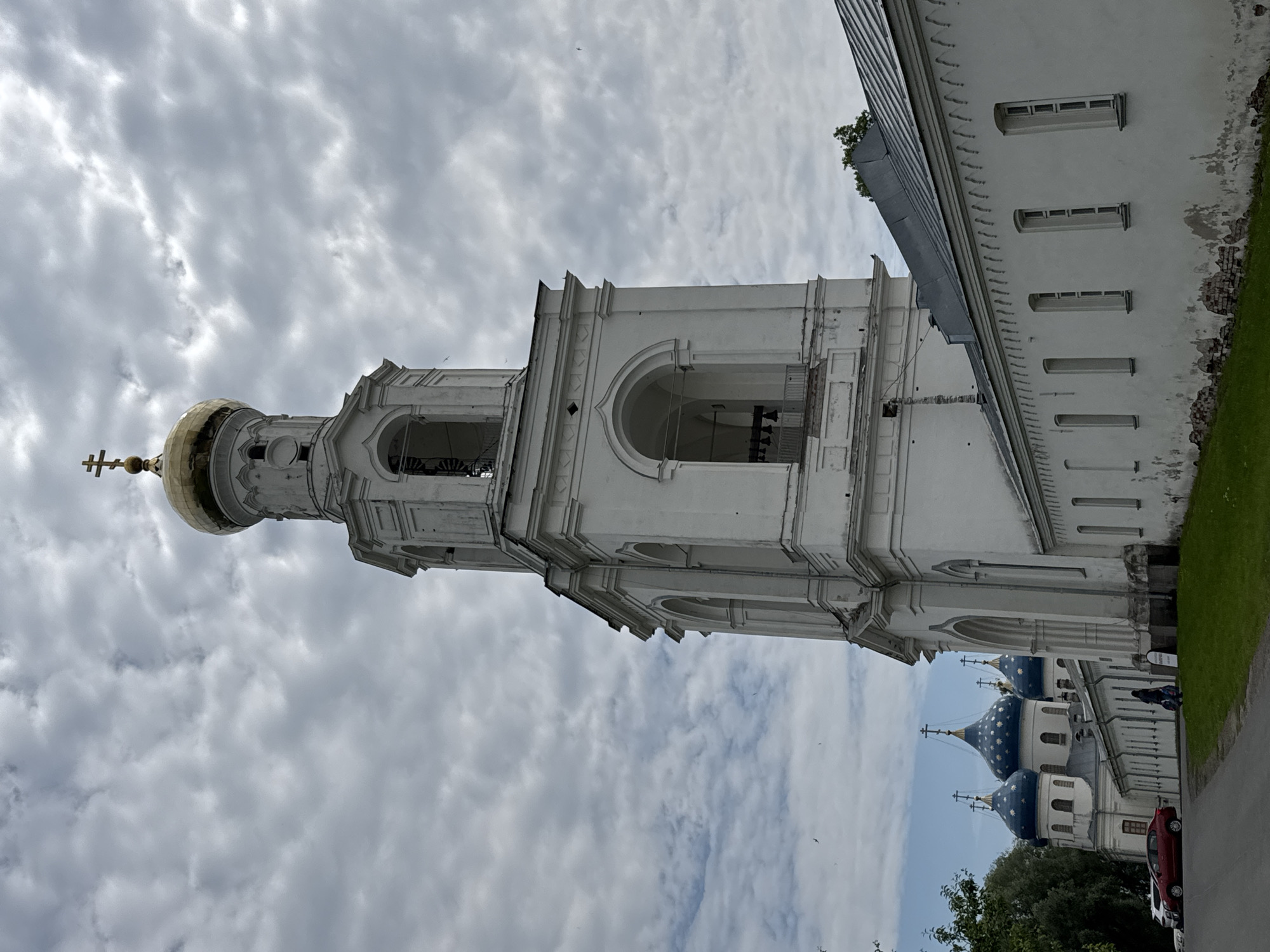 The image shows a white bell tower with a golden dome and a cross at the top, part of the Saint Sophia Cathedral complex in Veliky Novgorod, Russia. To the left of the bell tower, several blue domes with stars are visible on other church buildings. A long white wall with arched windows stretches along the foreground, with some signs of wear and tear at the base, revealing bricks. A red car is parked on the left side of the paved area in front of the wall. In the background, a few people are walking. The sky is overcast with dramatic clouds.