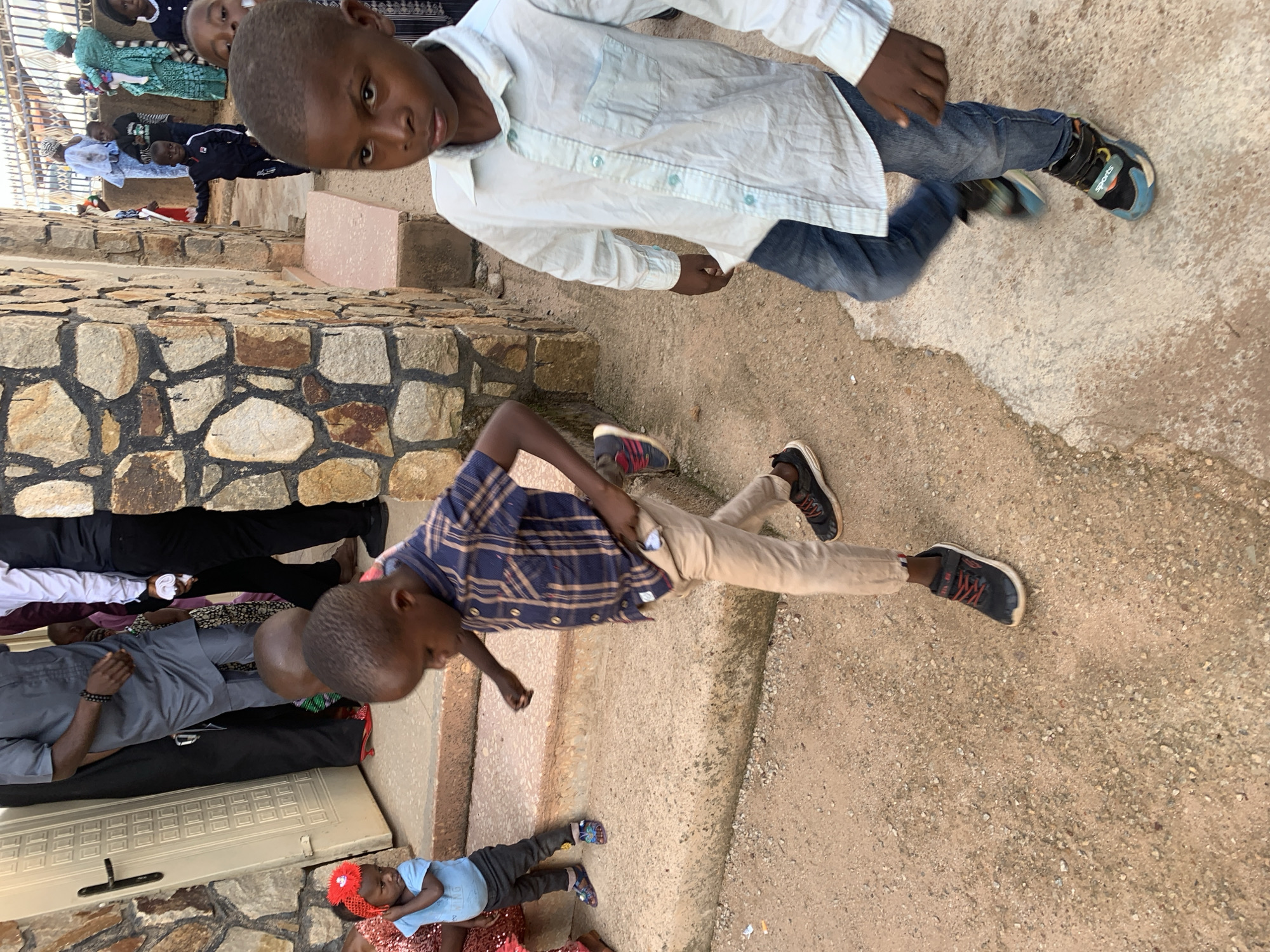The image depicts a group of people in Jos, Nigeria. In the foreground, two children are positioned on a concrete surface. The first child, wearing a white button-down shirt and blue jeans, is situated at the top. The second child, dressed in a blue plaid shirt and khaki pants, is lying below. Further back, a row of individuals is seen, resting on what appears to be a covered floor. A baby with a red bow in her hair is also visible at the end of the row. A stone wall borders the left side of the image. The environment is sunny, suggesting daytime.