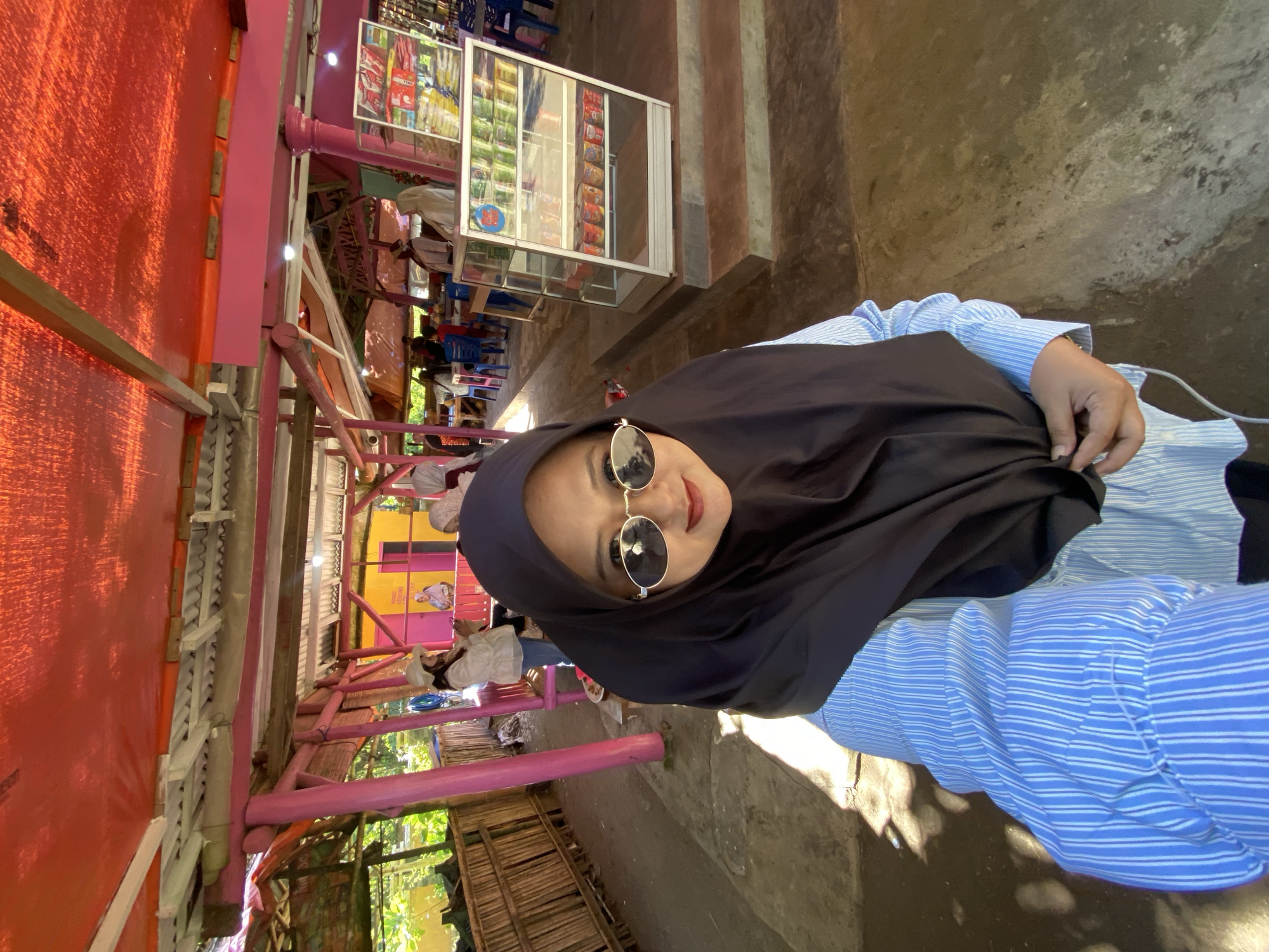 A woman wearing a black hijab and round sunglasses is posing for a selfie. She is wearing a blue and white striped shirt. Behind her is an outdoor space with pink posts and beams, and some bamboo structures. There is also a glass display case filled with goods. Several people are seated in the background. The location is in Indonesia, but the specific city is unknown.