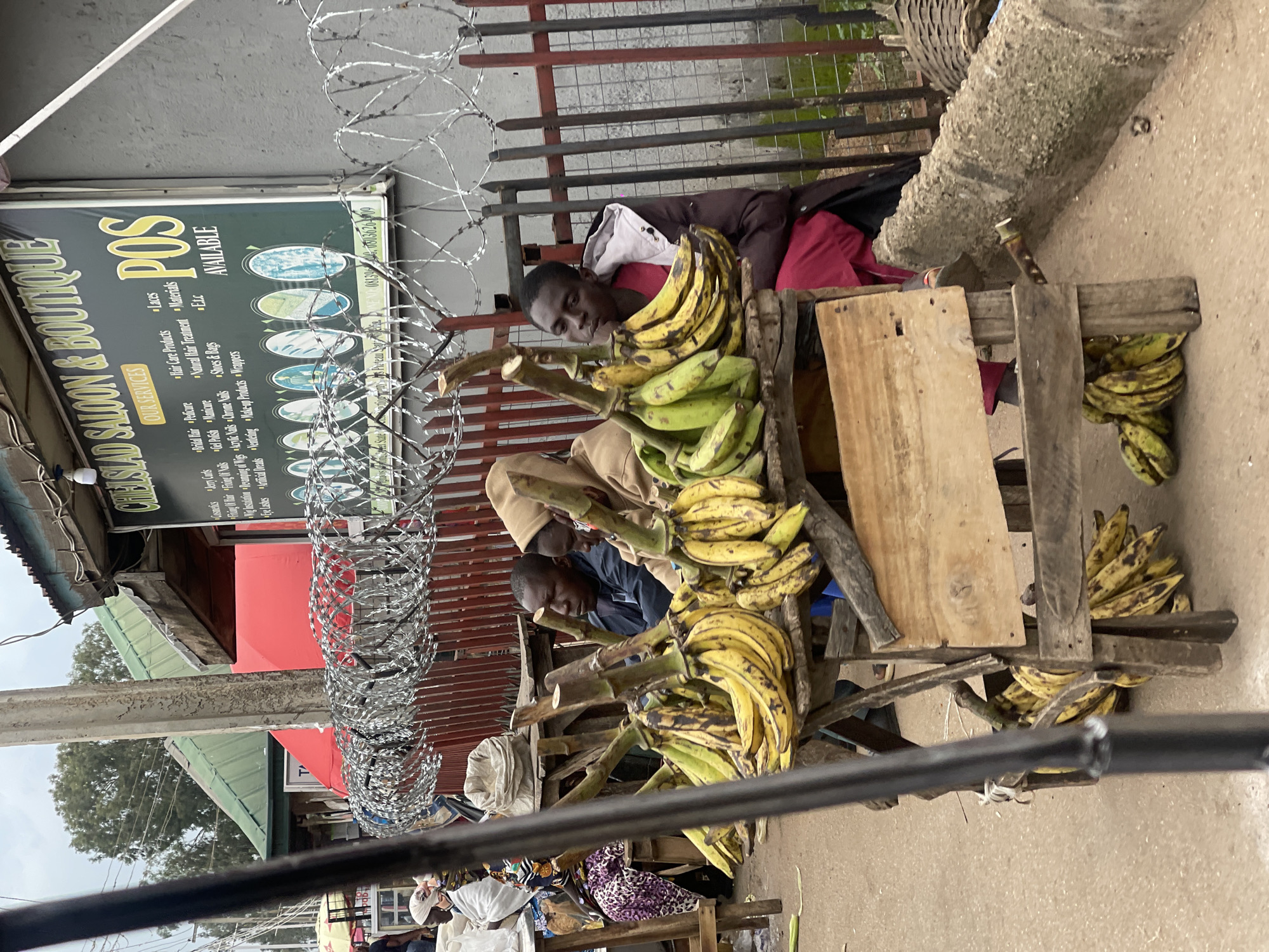 The image captures a street scene in Jos, Nigeria, where several individuals are engaged in the sale of bananas. Two men, one wearing a dark jacket and the other a light brown hooded jacket, are seated behind a display of bananas on a wooden stall. A third person, wearing a dark jacket and a bright pink garment, is also present near the bananas. In the background, a sign for "CHELSID SALOON & BOUTIQUE" is visible, advertising services such as hair care, manicure, pedicure, and the availability of POS services. Barbed wire fencing runs along the front of the scene, separating the market area from a road. Several bunches of yellow and green bananas are prominently displayed on wooden crates. The overall environment appears to be an outdoor market on a cloudy day, with buildings and trees visible in the distance.