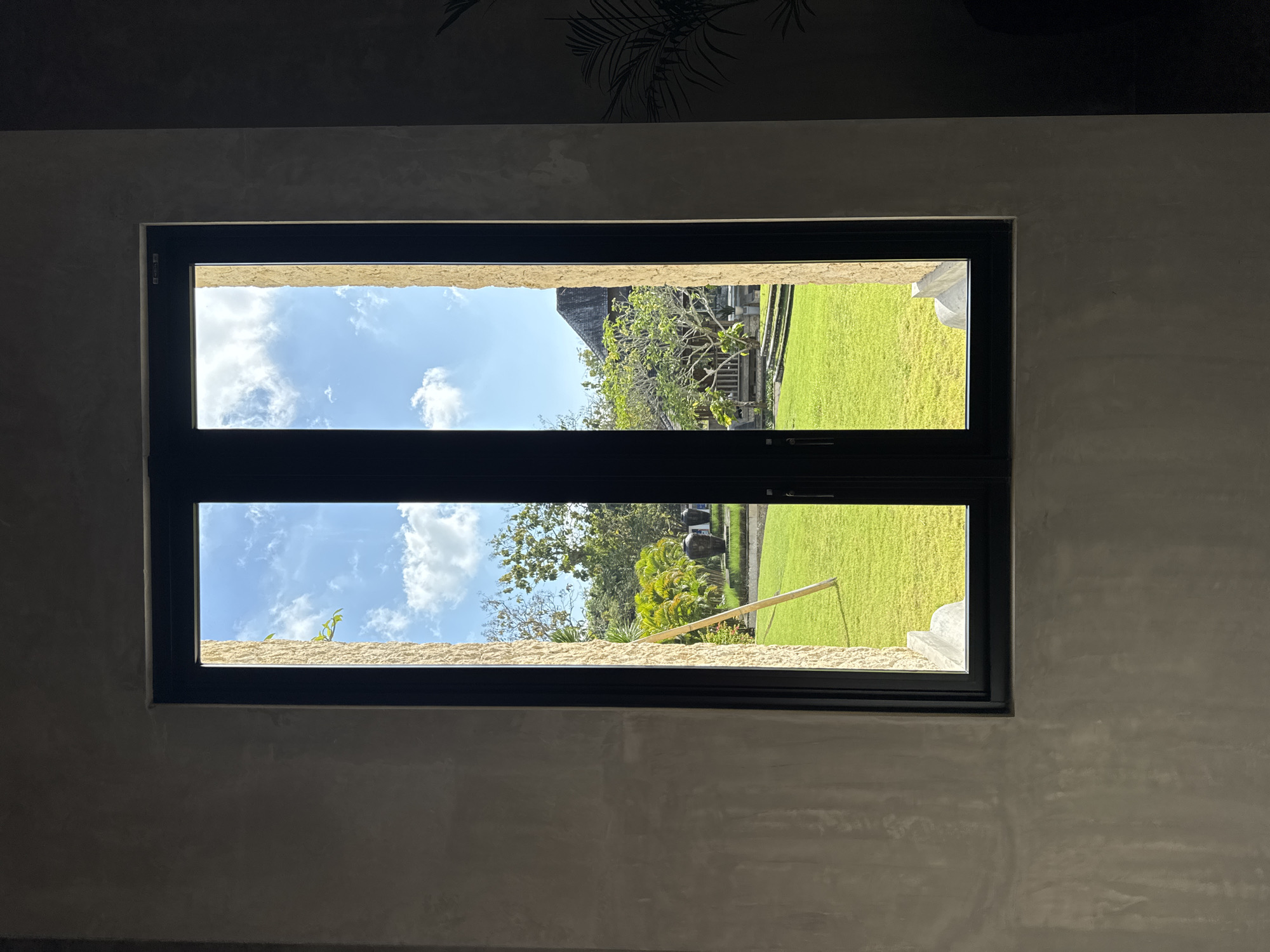 The image shows a view through a tall, narrow window with black framing, offering a glimpse of a sunlit outdoor scene in Kuta Selatan, Indonesia. The window is divided vertically into two panes. Through the left pane, the bright blue sky with scattered white clouds is visible, along with some green foliage. The right pane reveals more of the outdoor environment, including a lush, well-maintained green lawn.
Dominating the outdoor view is a large, mature tree with a spreading canopy, positioned towards the right side of the frame. In front of the tree, there are stone steps leading up to a wooden structure that appears to be part of a building or pavilion. To the left of the tree, a pale wooden pole is propped diagonally against the lawn, possibly for support or decoration. In the background, beyond the lawn and tree, there are more trees and hints of other structures, including a dark-roofed building that resembles a traditional house. Two large, dark urns are visible on the lawn, placed in front of a row of smaller plants.
The interior framing of the window has a rough, textured surface, possibly concrete or stucco, with variations in color and tone, suggesting it is part of an indoor wall. The exterior framing of the window appears to be a light-colored stone or stucco. The overall lighting suggests it is daytime, with the sun casting shadows and illuminating the outdoor landscape.