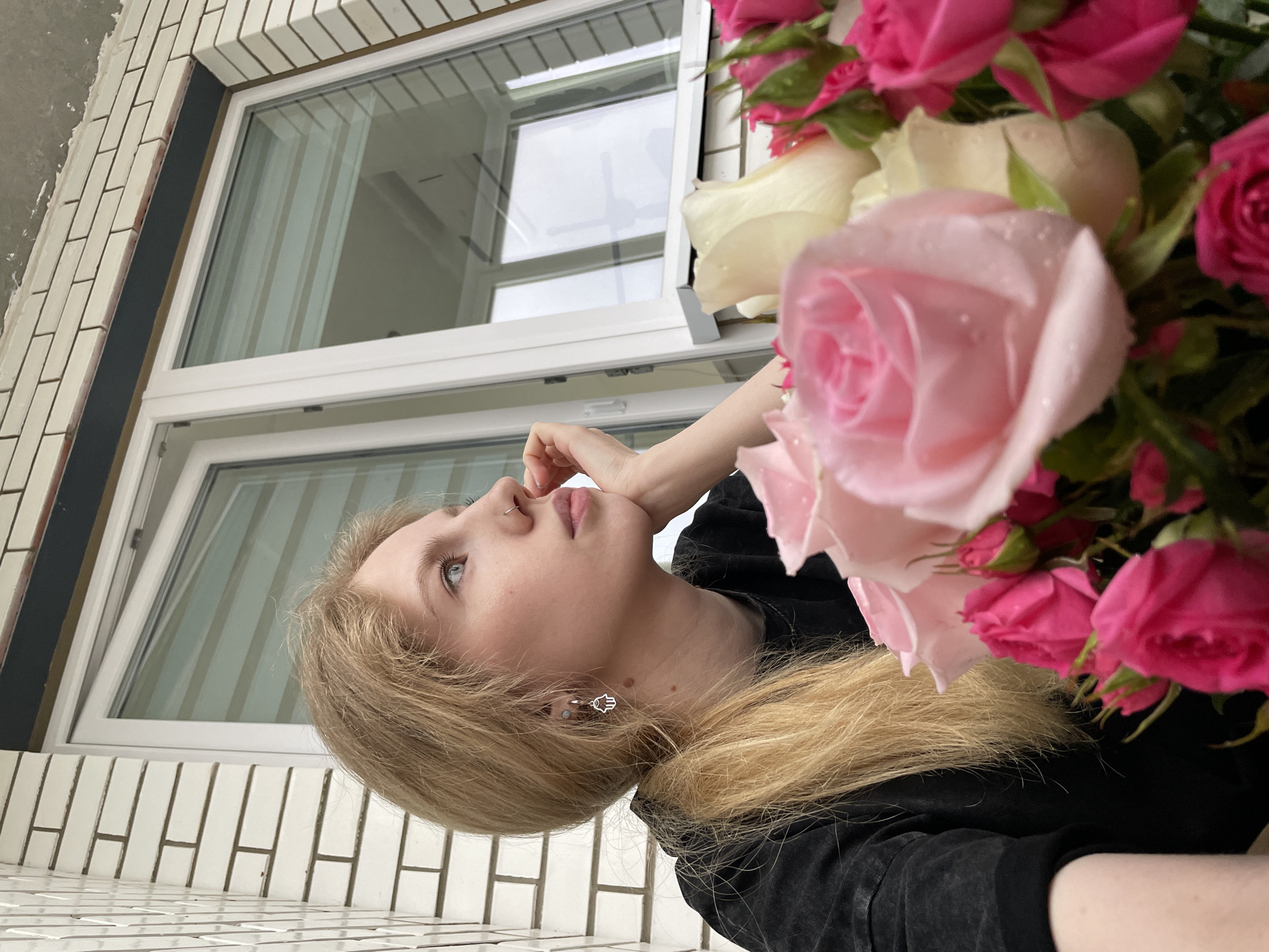 A photograph shows a blonde woman with blue eyes standing on a balcony with a brick railing and a window behind her. She is wearing a black top, and her head is tilted upward, her hand near her face. A bouquet of pink and white roses is positioned to the right, partially obscuring her. The window reflects the sky. The location is Izhevsk, Russia, indicated by the architectural style of the building and window.