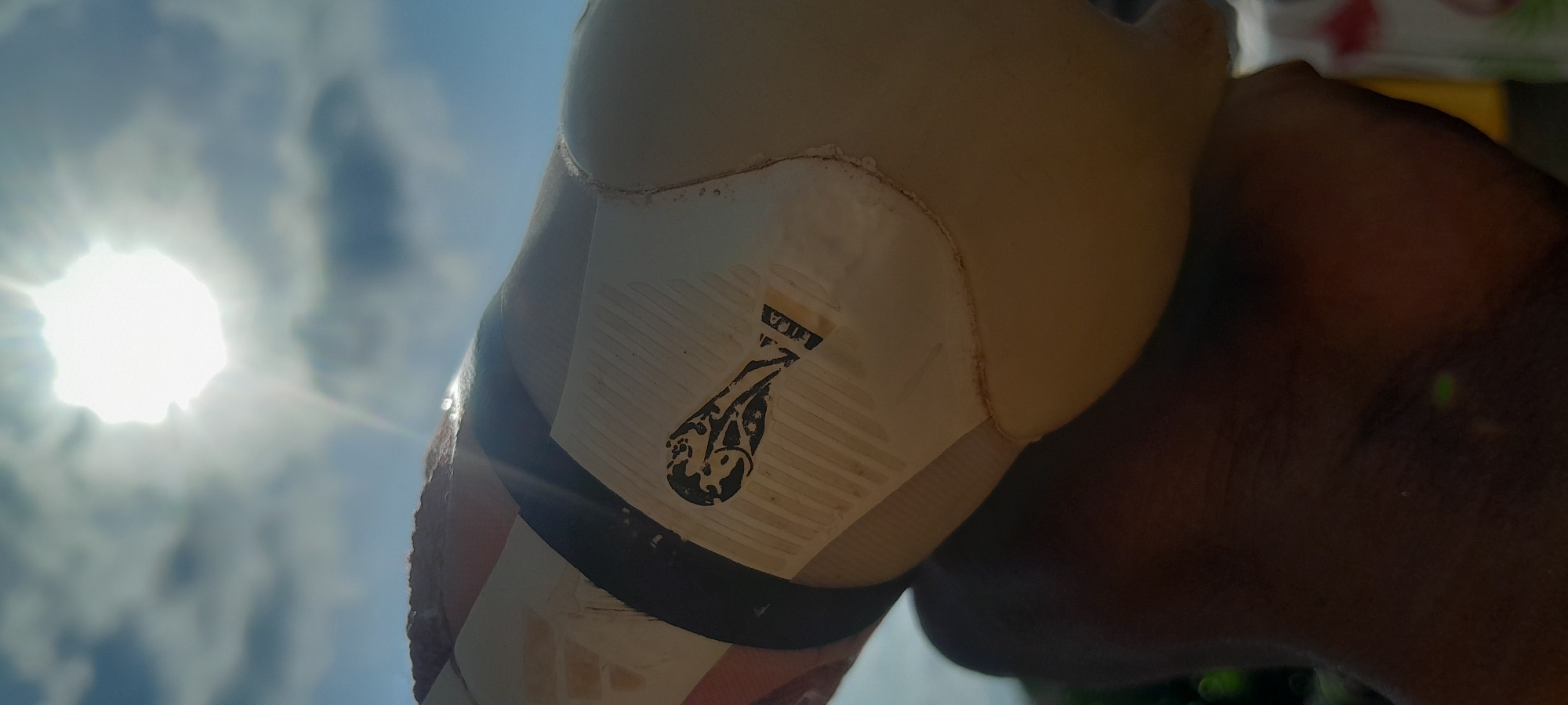 An extreme close-up, low-angle shot captures a partially deflated, beige-colored soccer ball held by a dark-skinned hand. The ball, worn and slightly misshapen, displays a black FIFA logo and branding with text. Sunlight beams intensely from a bright, overexposed sky filled with scattered clouds, creating a lens flare effect and casting a bright glow at the top center of the frame. The background consists of a blue sky with clouds and hints of greenery at the bottom left, suggesting an outdoor environment. The image is underexposed, with the bottom half of the frame showing the dark skin of the hand and the dark shadows of the ball. The scene is set in Abuja, Nigeria, likely during daylight hours.