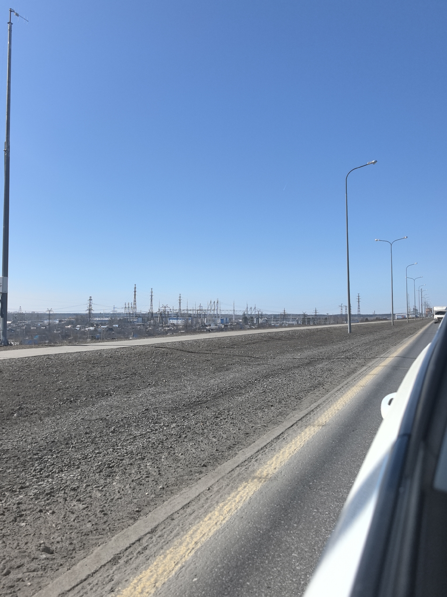 The image shows a road in Chaikovsky, Russia, with a view of an industrial area in the distance. The road has a wide, unpaved shoulder covered in gray gravel. A white vehicle is partially visible on the right side of the frame. Streetlights line the right side of the road. In the background, a cityscape with power lines and industrial towers is visible under a clear blue sky. A tall metal pole is on the left of the frame.