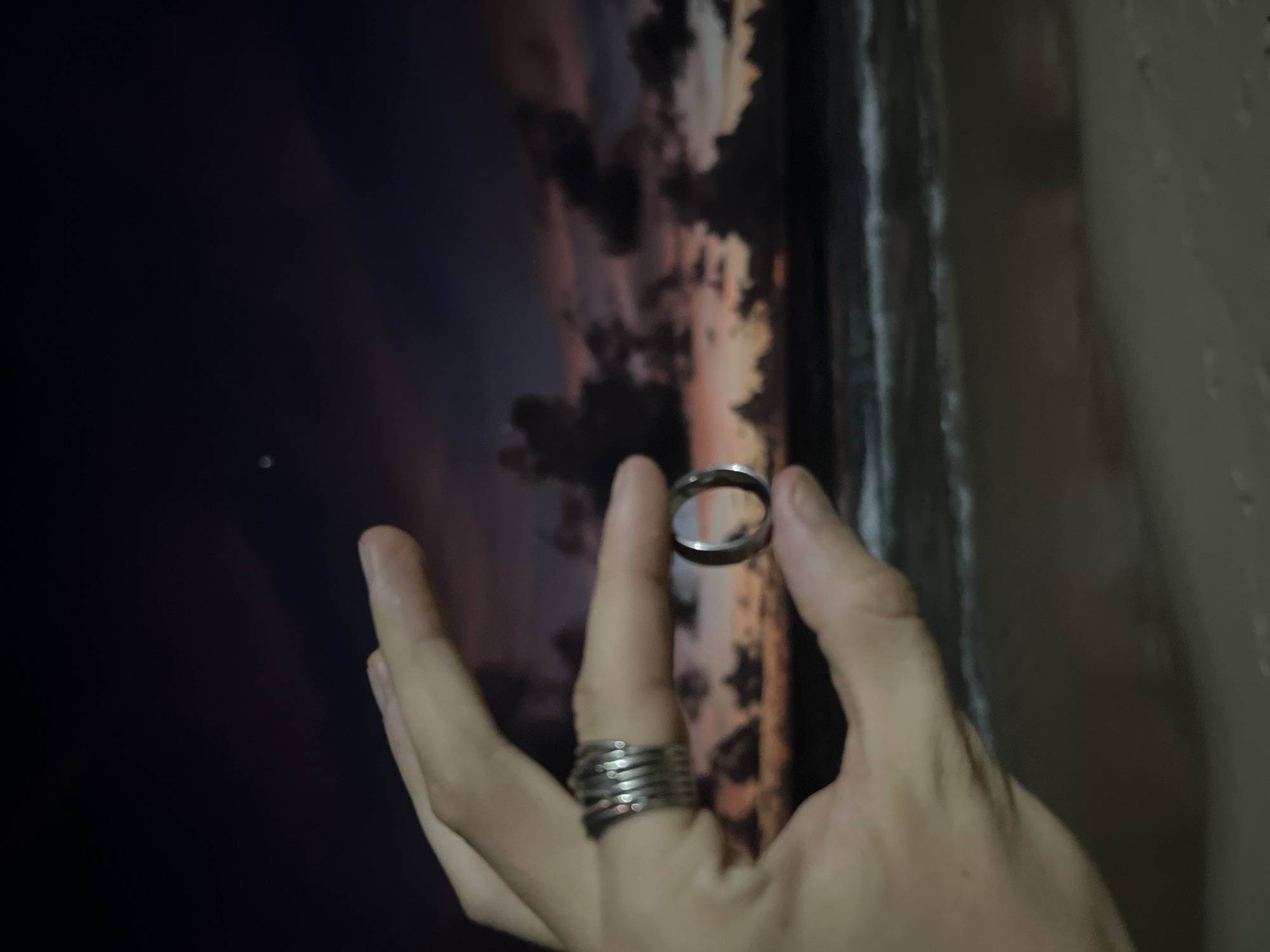 The image shows a close-up of a person's hand holding two rings. One ring is on the person's index finger, and it appears to be a stacked, silver ring with a coiled design. The other ring is being held between the thumb and index finger of the same hand, and it is a simple, silver band. The background is blurred and appears to be a wall with a picture on it. The picture is of a dark sky with colorful clouds, possibly a sunset or sunrise. There is a hint of a vertical, dark line on the right side of the image, which could be a window frame or a door. The lighting suggests it is either dusk or dawn, or the room is dimly lit. The person's hand is pale, and the overall tone of the image is muted. There is no visible text in the image.