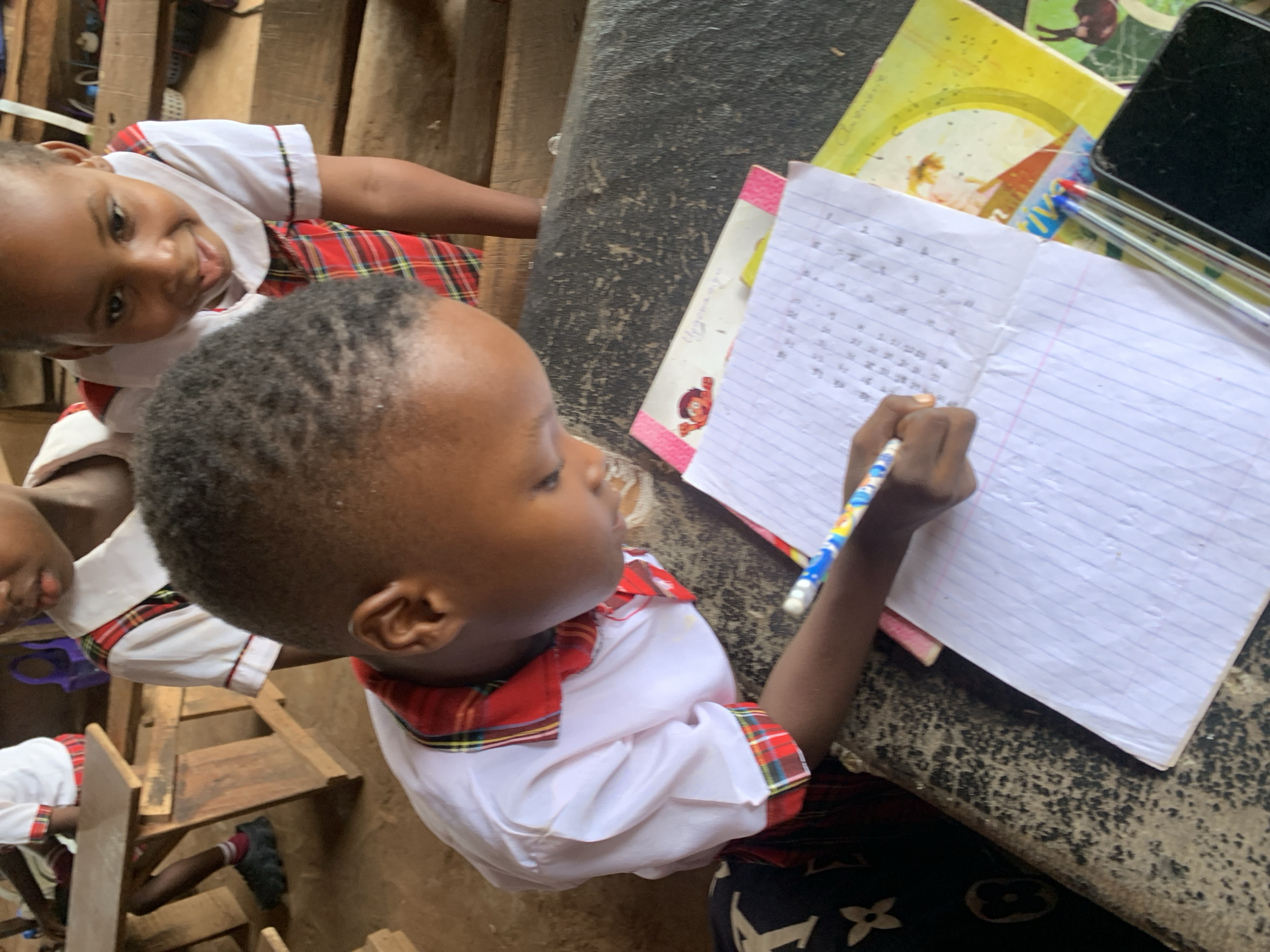 In Ukwu-Abwa, Nigeria, the image depicts two children seated at a worn desk. One child is actively writing in a notebook. The other child watches, leaning close. The writing child wears a white shirt with a red and green plaid collar and sleeves, paired with plaid shorts. A notebook, a yellow book, pens, and a mobile phone are arranged on the desk. The environment around the desk appears to be simple, possibly a classroom or home setting. Wooden furniture is visible in the background.