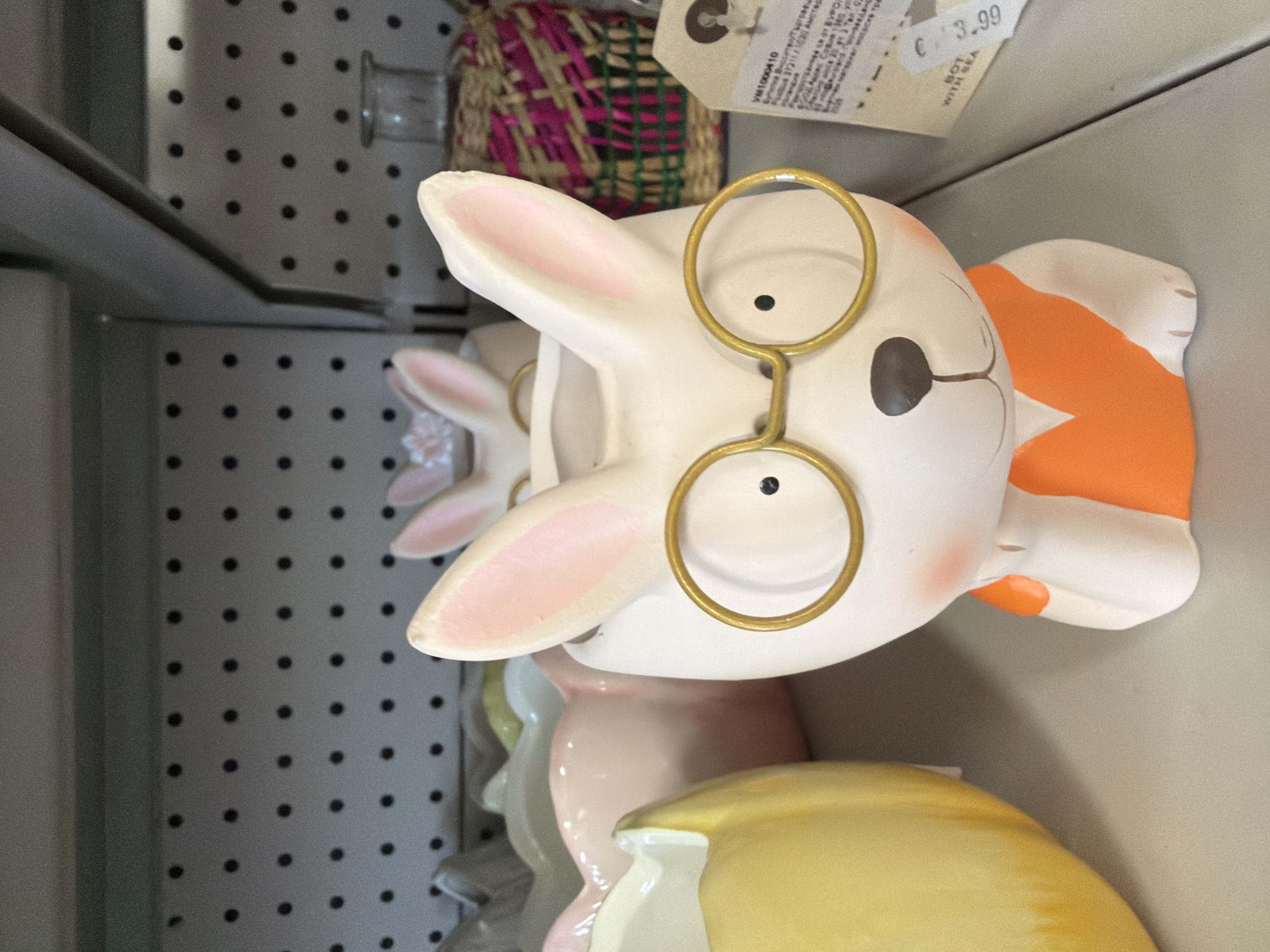A white ceramic bunny planter with pink-lined ears and gold-rimmed glasses sits on a shelf. The planter is shaped like a bunny's head and torso, with an orange V-neck shirt design. The bunny's right hand is positioned near its cheek, as if resting its head. Behind the bunny, other planters in various pastel colors are visible. To the right of the bunny planter, a woven basket with pink and green accents and a small glass bottle can be seen. A price tag displaying "€ 3.99" is attached to the right side of the image. The background consists of a gray pegboard. The location is a retail environment, likely a store, and no specific city in Pazardzhik, Bulgaria is identifiable.