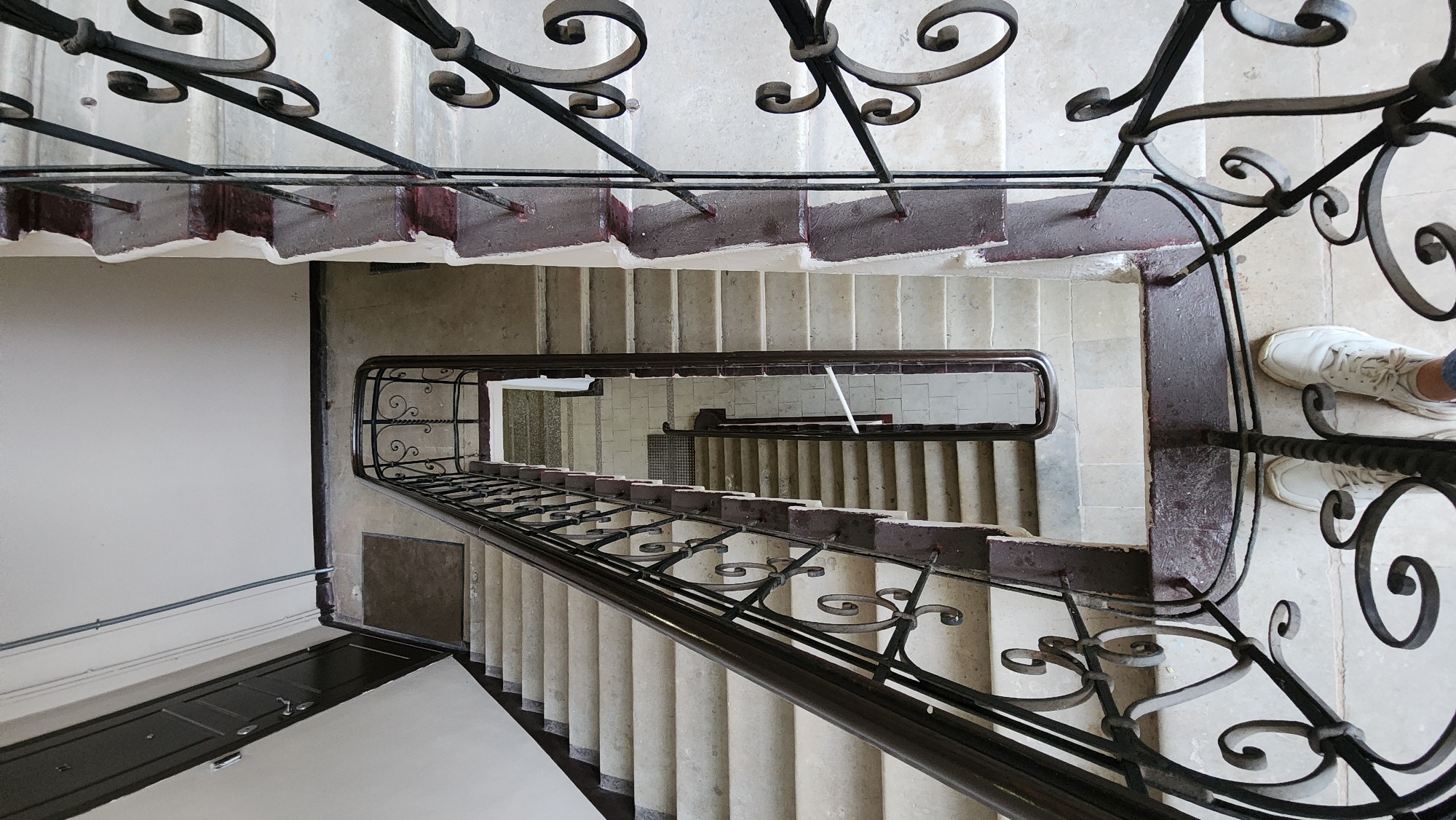 A high-angle shot shows a winding staircase with an ornate wrought-iron railing. The steps are light-colored stone or concrete with dark brown edging. The walls are painted white, and a dark door is visible on the left side of the image. A pair of white sneakers is visible at the right edge of the frame. The location is unknown.