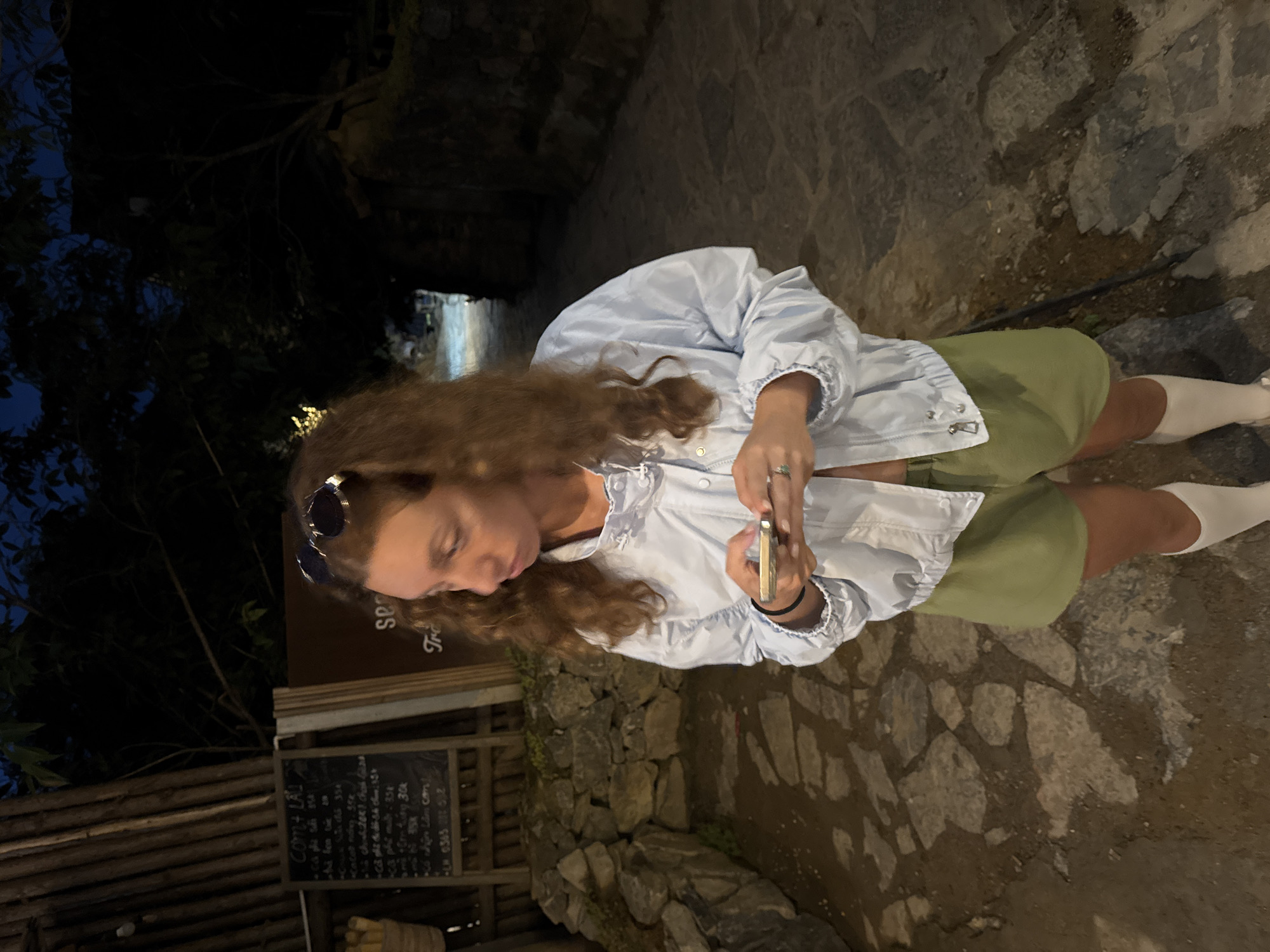 The image depicts a young woman with long, curly brown hair standing outdoors in what appears to be Lũng Cú Commune, Vietnam. She is wearing sunglasses perched on her head, a light blue ruffled jacket, green shorts, and white socks. She is holding a smartphone in her hands and looking down at it, possibly interacting with the device.
The background consists of a stone pathway, a wall constructed of similar stones, and wooden structures. A menu board with Vietnamese writing is visible near her, suggesting the setting might be near a local establishment. The scene is dimly lit, likely indicating that the photo was taken at dusk or during the evening.