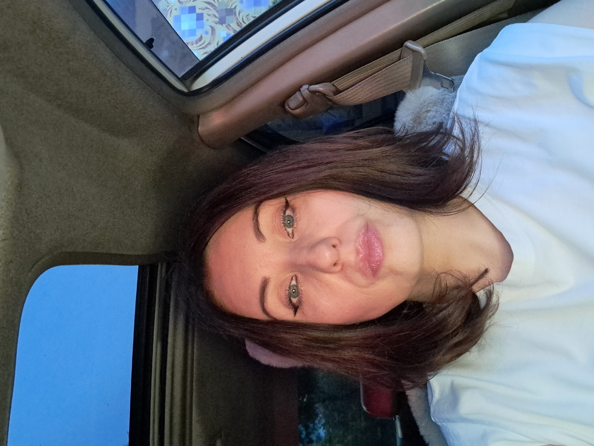 A young woman with dark hair and light blue eyes is taking a selfie inside a vehicle. She is wearing a white t-shirt and has eyeliner and lip gloss on. The sun/moon roof of the car is open, revealing a clear blue sky. The interior of the car is visible, including a beige headliner, a brown seatbelt, and a red car seat. The background outside the car window shows trees and foliage, indicating an outdoor setting, potentially in Irkutsk, Russia.