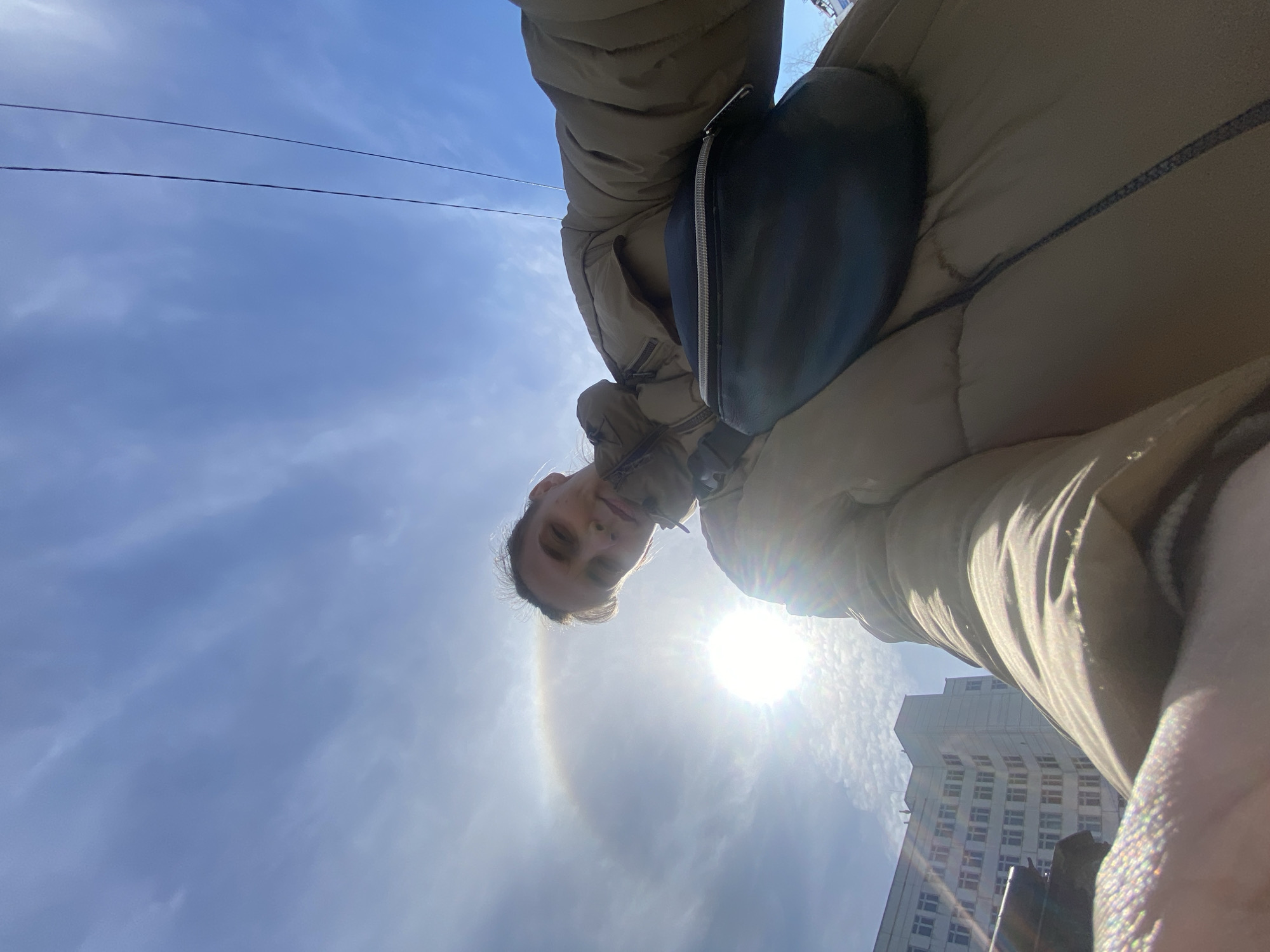 A low-angle, rotated selfie shows a person wearing a puffy beige jacket and a black crossbody bag. The background features a bright, sunlit sky with a faint halo effect around the sun. In the lower right corner, a portion of a tall, white building is visible. Two thin, dark power lines extend across the upper left portion of the sky. Based on the building architecture and subject location, the image was captured in Chaikovsky, Russia.