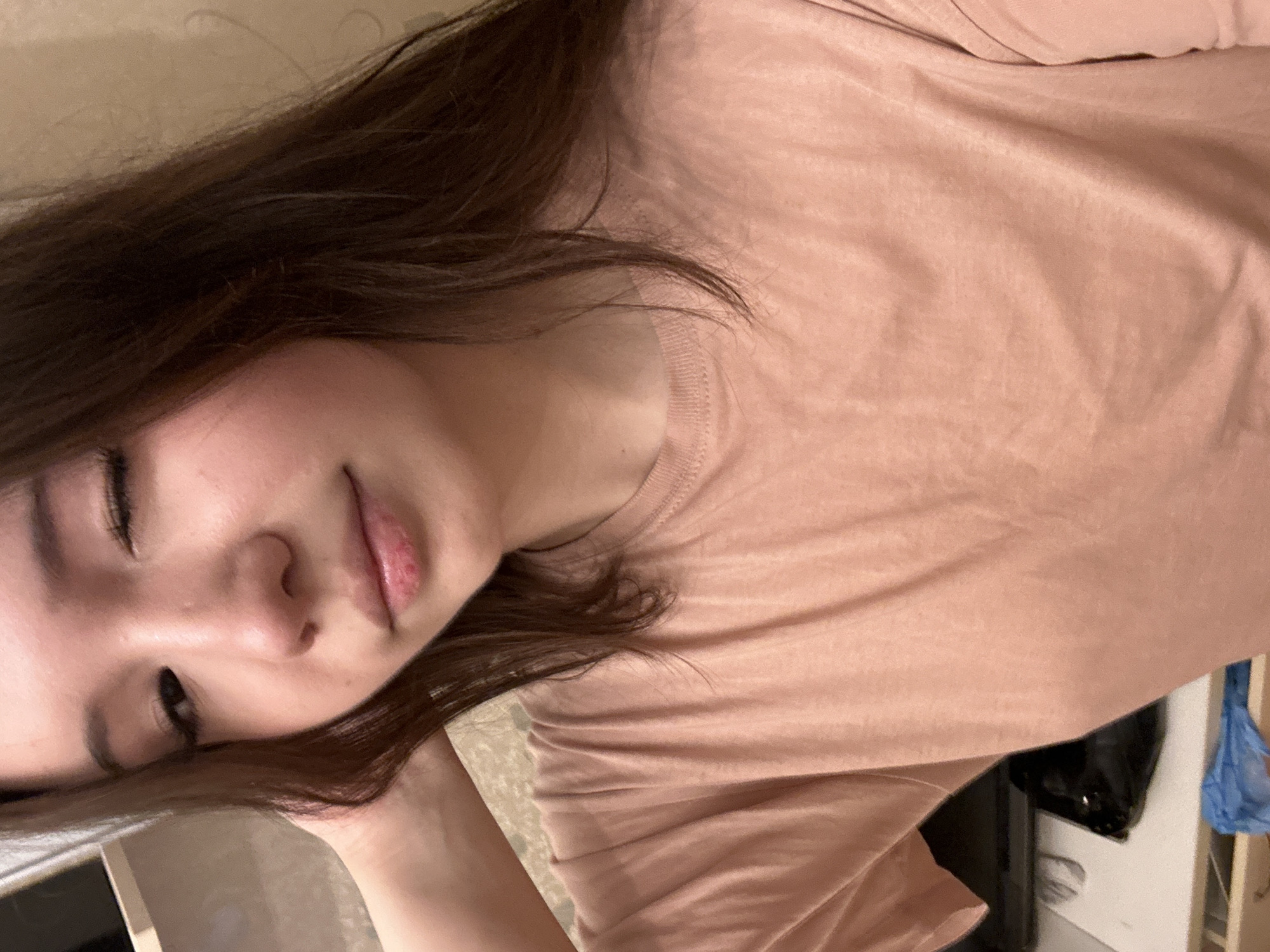 This close-up portrait, taken in Naberezhnye Chelny, Russia, features a young woman with dark brown, long hair, appearing to be resting or relaxing indoors. Her head is tilted, suggesting she is lying down or leaning back. She is wearing a simple, light peach or dusty pink crew-neck t-shirt made of a soft, slightly wrinkled fabric.
The woman's eyes are closed or mostly closed, and her lips are slightly parted, revealing a soft, natural lip color. Her facial expression is peaceful and serene, conveying a sense of calm or sleepiness. The lighting in the image is soft and warm, indicative of an indoor setting, possibly under artificial light or during the evening.
The background elements are largely out of focus, emphasizing the subject. Hints of a casual, domestic environment are visible. To the upper right, a light-colored wall or surface is present. Below the edge of her shirt on the right side, a dark, rectangular object, possibly an electronic device, and a crumpled blue plastic bag can be seen. A light-colored object is also partially visible on the far left, near her head. There is no visible text in the image.
