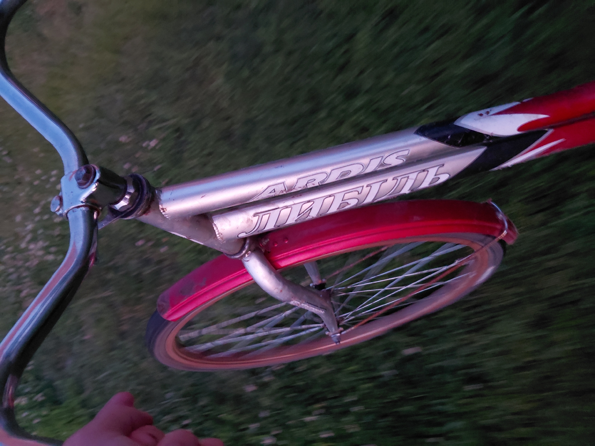 A first-person perspective shot of a bicycle with a red fender is captured. The rider's hand is visible on the left side of the image, and the bicycle frame, which is silver with red and white decals, is prominent. The front wheel of the bicycle is in focus, showing its spokes and rim. The background consists of blurred green grass, suggesting the bicycle is in motion. The scene is set outdoors in Ustyvytsia, Ukraine.