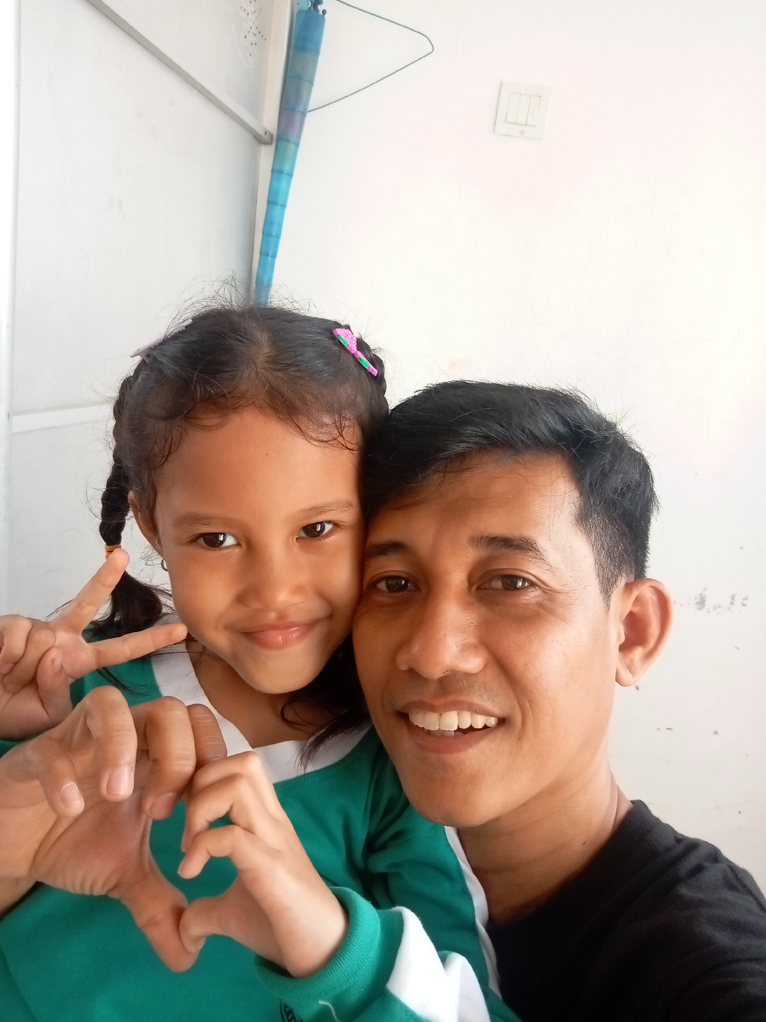 A medium shot shows a man and a young girl posing for a selfie. The man is on the right, a fair-skinned man with short black hair and a slight smile. He is wearing a black t-shirt. The girl is on the left, a fair-skinned girl with dark hair in two braids, with pink and green hair clips. She is wearing a green and white shirt. She is smiling and making a peace sign with her left hand, and her right hand is forming a heart shape with her left hand. The background is a plain white wall with a light switch and a blue tube-like object hanging from the ceiling. The scene appears to be indoors, possibly in a home in Batam, Indonesia.