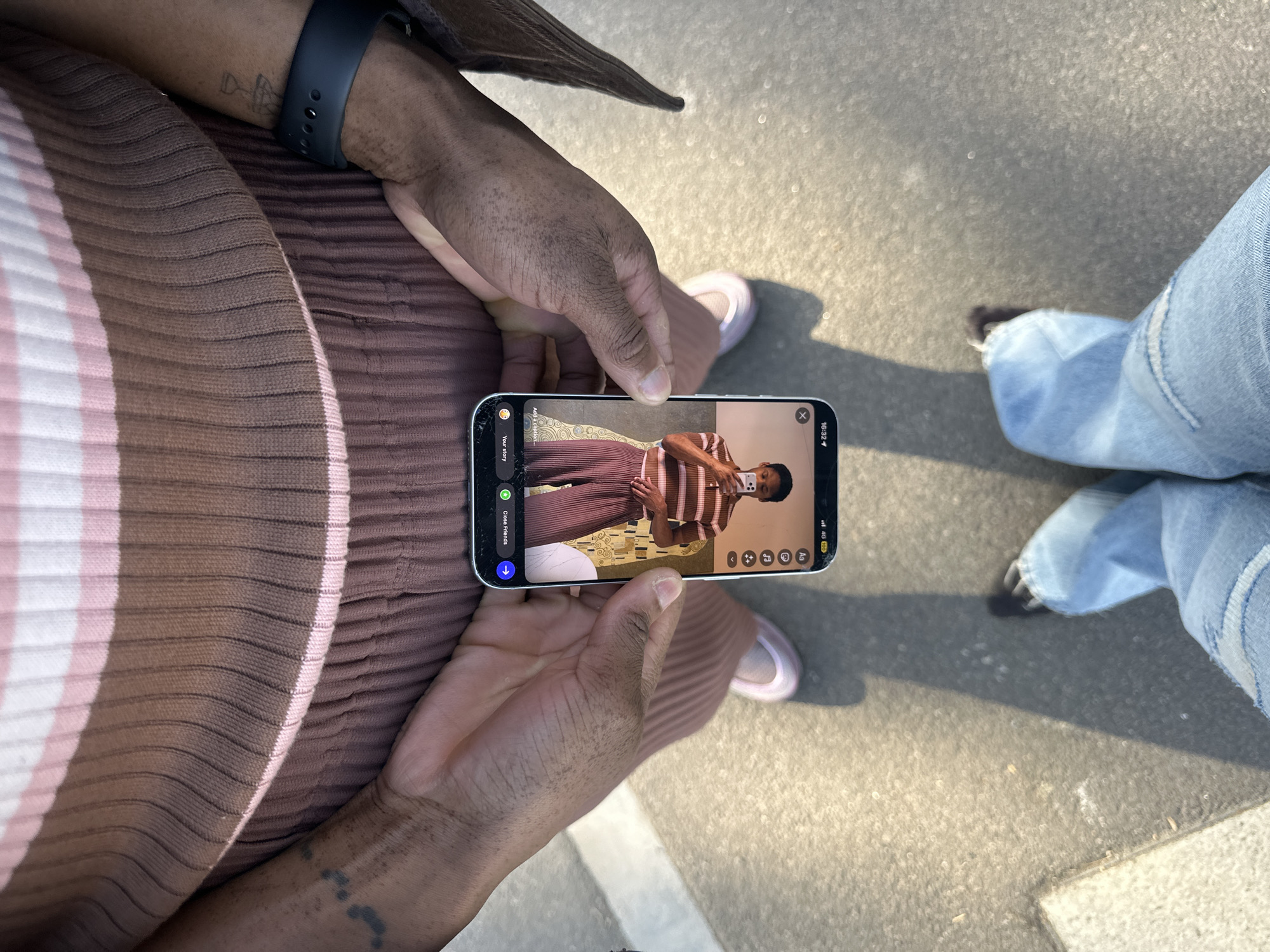 The image shows a person holding an iPhone displaying an image of themselves. The person holding the phone is wearing a dark-colored smart watch and a striped brown and pink top. They are also wearing pink sneakers. The iPhone's screen displays the person in a tan top with brown stripes and matching brown pleated pants. They appear to be taking a selfie in front of a decorative gold-colored screen or wall. The background shows a cement surface. To the right of the person with the phone are legs clad in blue jeans. The location appears to be outdoors. There is no information to specify this image was taken within Folkestone and Hythe District, United Kingdom.