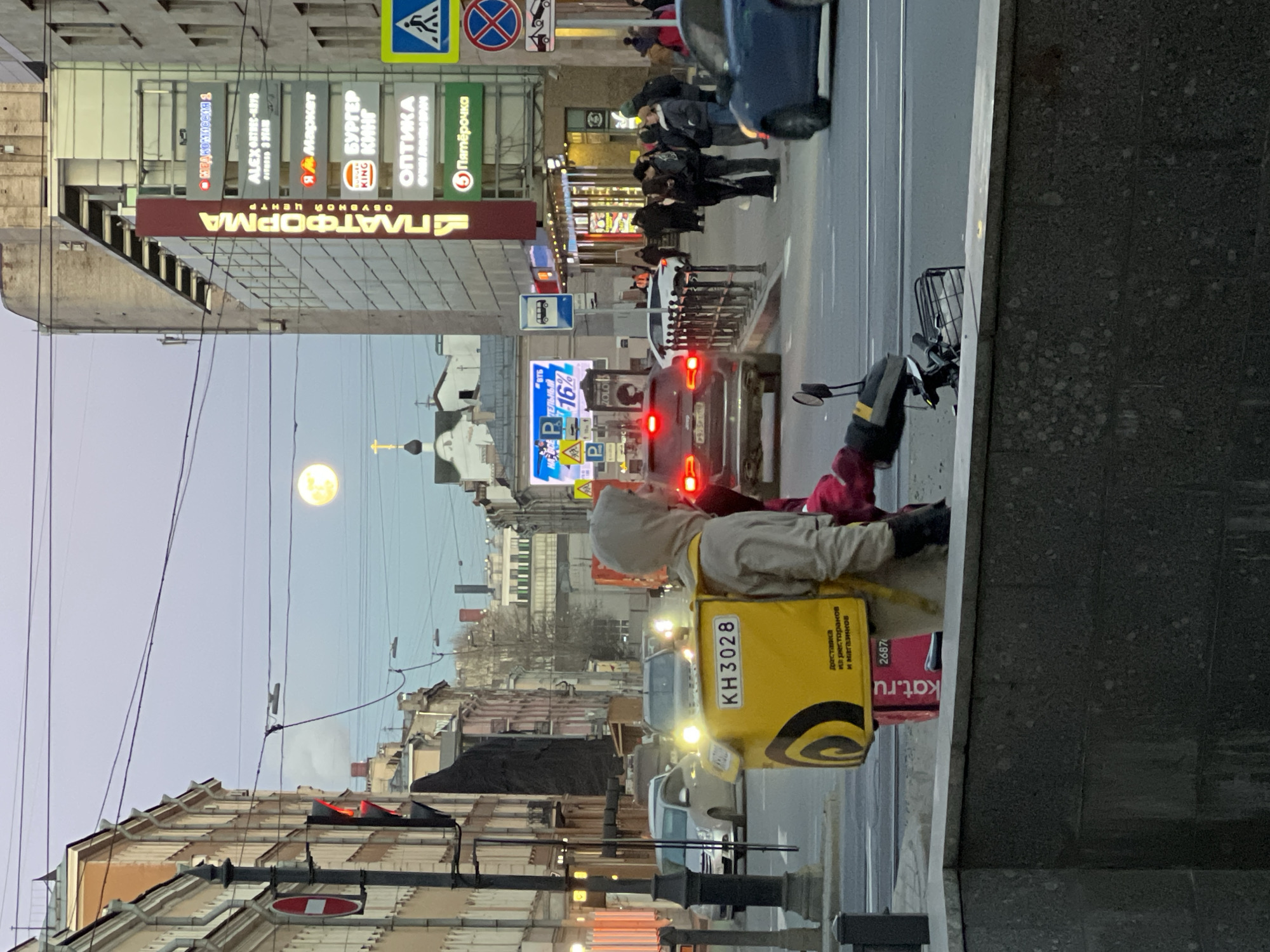 The image captures an urban scene in Saint Petersburg, Russia, at dusk or dawn, with a clear sky and a bright moon visible. In the foreground, a delivery person wearing a tan outfit and a yellow "Delivery Club" branded backpack is stationary on a scooter. The backpack has the license plate "KH3028" and website "kat.ru". The rider is stationary near a sidewalk curb. Behind the scooter, a street with car traffic is visible. Across the street, commercial buildings house various establishments, including "Platforma" shopping center, Burger King, and a "Pyaterochka" store. A church with a golden dome and cross stands out in the background. Pedestrians are visible walking along the street. Street signs and advertisements are also present. The overall impression is a typical urban scene in Saint Petersburg, Russia.