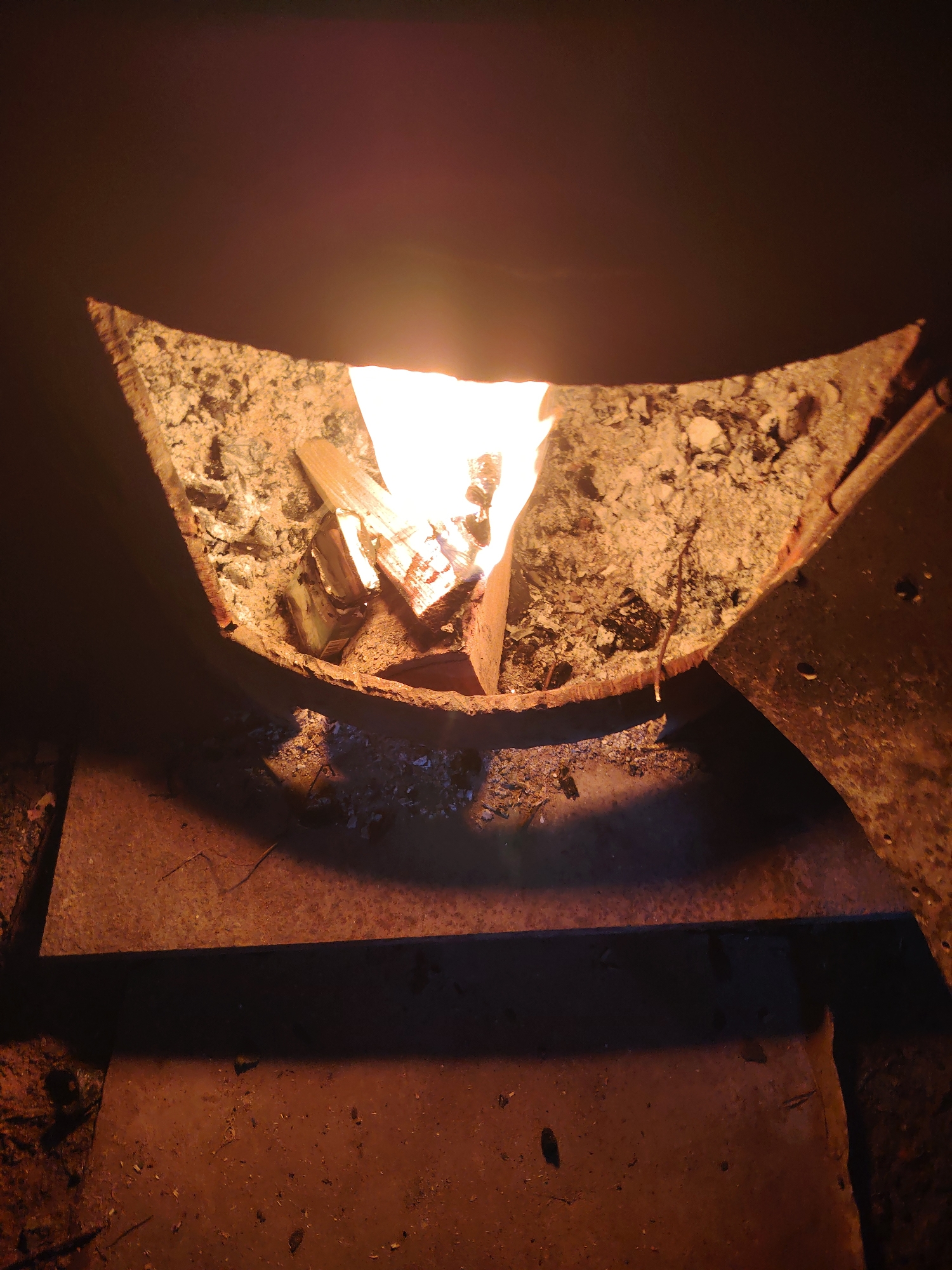 A high-angle, medium shot shows a fire burning inside a crude, open-topped fire pit constructed from dark, possibly metal, materials. The pit's shape resembles a wide "U". Ash and unburnt debris line the inside edges, surrounding a small fire burning near the pit's center. Below the pit, a dark, flat surface shows shadows cast by the pit and fire, suggesting an indoor environment. The overall lighting is dim and predominantly orange-toned due to the fire. Based on the image alone, it is impossible to determine the exact location or whether it is related to Chaikovsky, Russia. There are no visible people or animals in the frame.