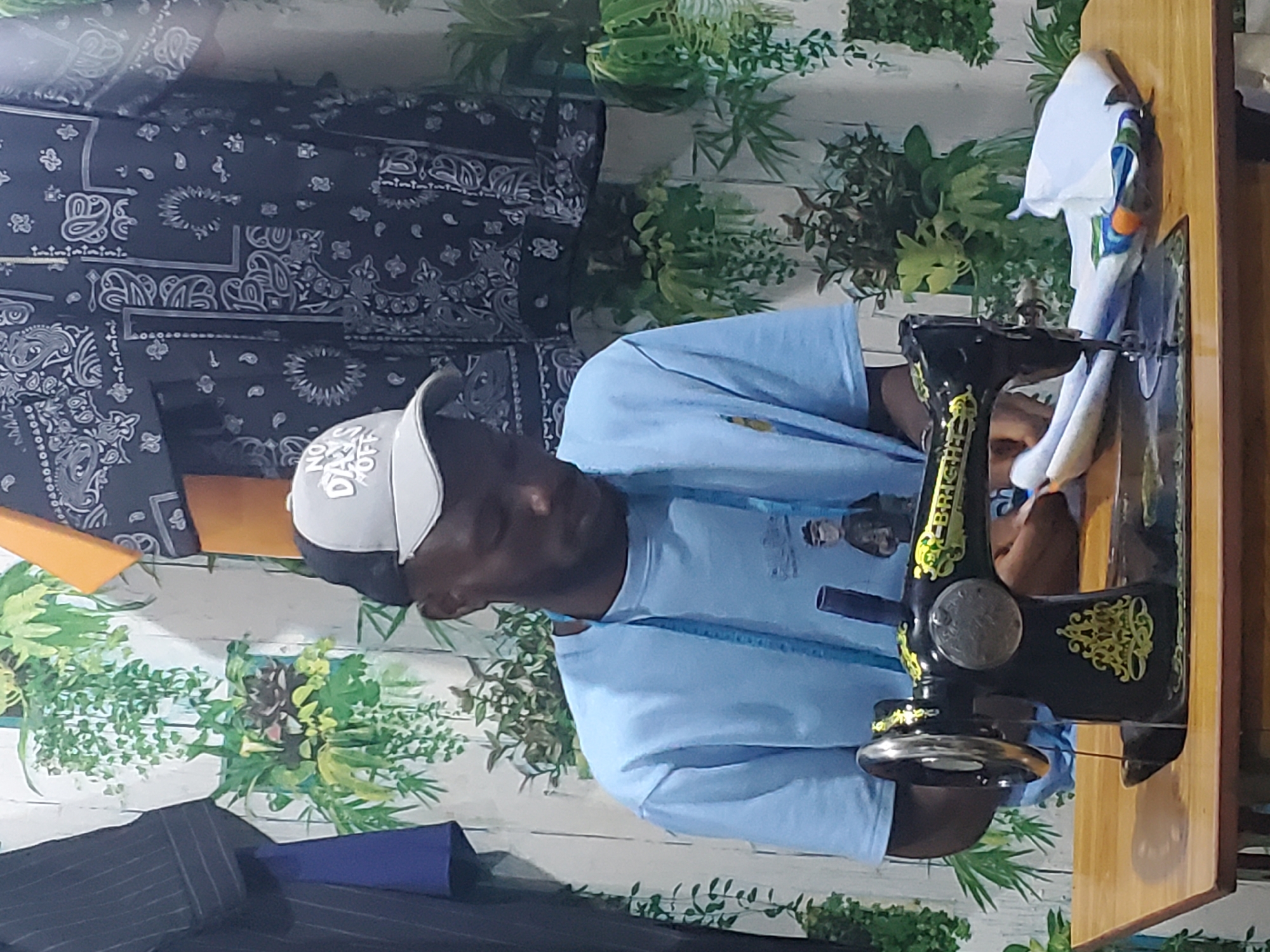 The image shows a man in a light blue t-shirt and a grey and black baseball cap working at a wooden table. He is seated and looking down at a black and gold sewing machine in front of him. A light blue measuring tape is draped around his neck. Behind him, the wall is decorated with a printed pattern of hanging plants. To his left, partially visible, is a dark pinstripe garment, and to his right, a dark fabric with a paisley pattern. The scene appears to be an indoor workshop or tailoring space in Jos, Nigeria.