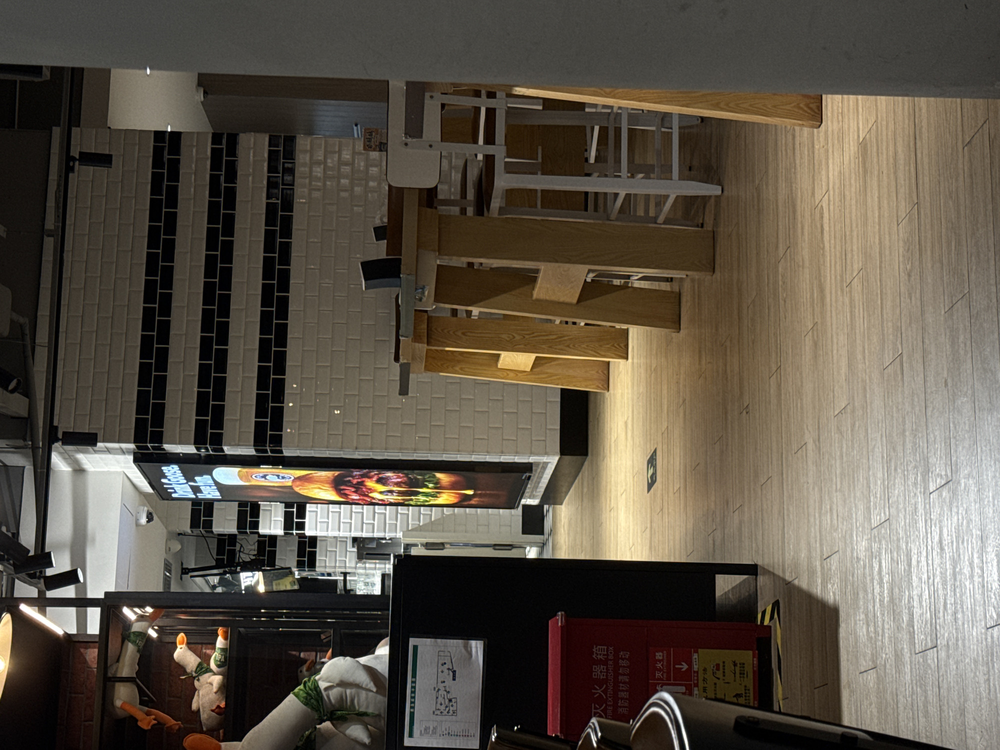 The image shows an interior scene, likely a restaurant or cafe, with a focus on the dining area. In the foreground, a black object, possibly a table or booth, is partially visible. To the left, a red fire extinguisher box is prominent, with Chinese text indicating it is a fire extinguisher. Above it, a diagram or map is mounted on a black surface. The floor is made of light-colored wooden planks.
In the mid-ground, several tall wooden tables are set up with white stools, suggesting a bar or high-top dining area. Behind the tables, a tiled wall with horizontal black stripes forms a backdrop. A large illuminated sign with a picture of a burger and text is displayed vertically on this wall. The text on the sign includes "Drink Goose" and "Have Fun."
In the background, shelves are visible displaying stuffed animal toys, some resembling ducks with green scarves. The overall lighting is dim, with bright accent lights highlighting certain areas. The scene suggests a casual dining establishment. The provided context states the location is Pudong, China.